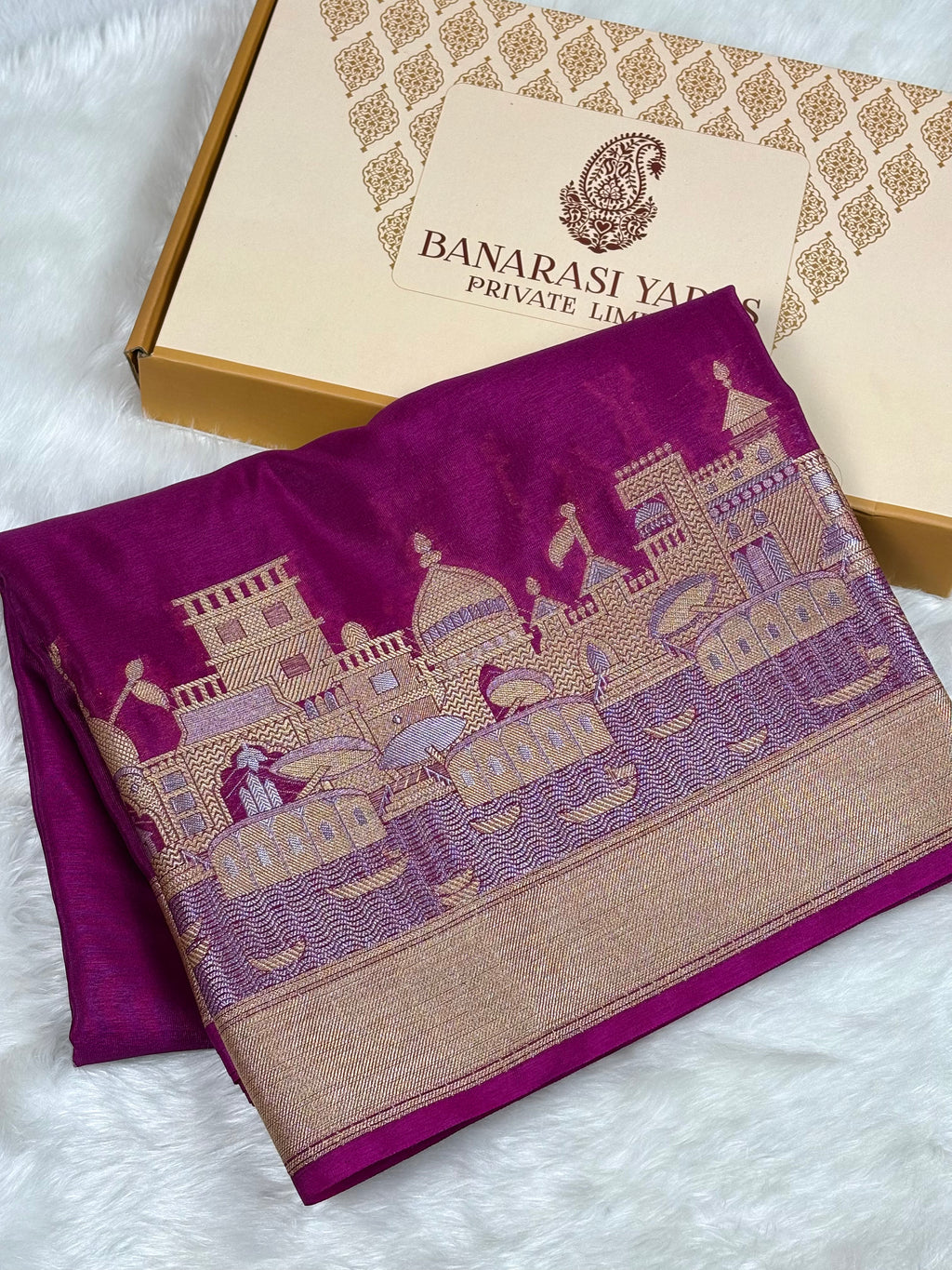 A folded magenta purple Banarasi silk saree with a detailed alifi mina zari architectural border, displayed next to its branded cardboard box on a white faux fur surface.