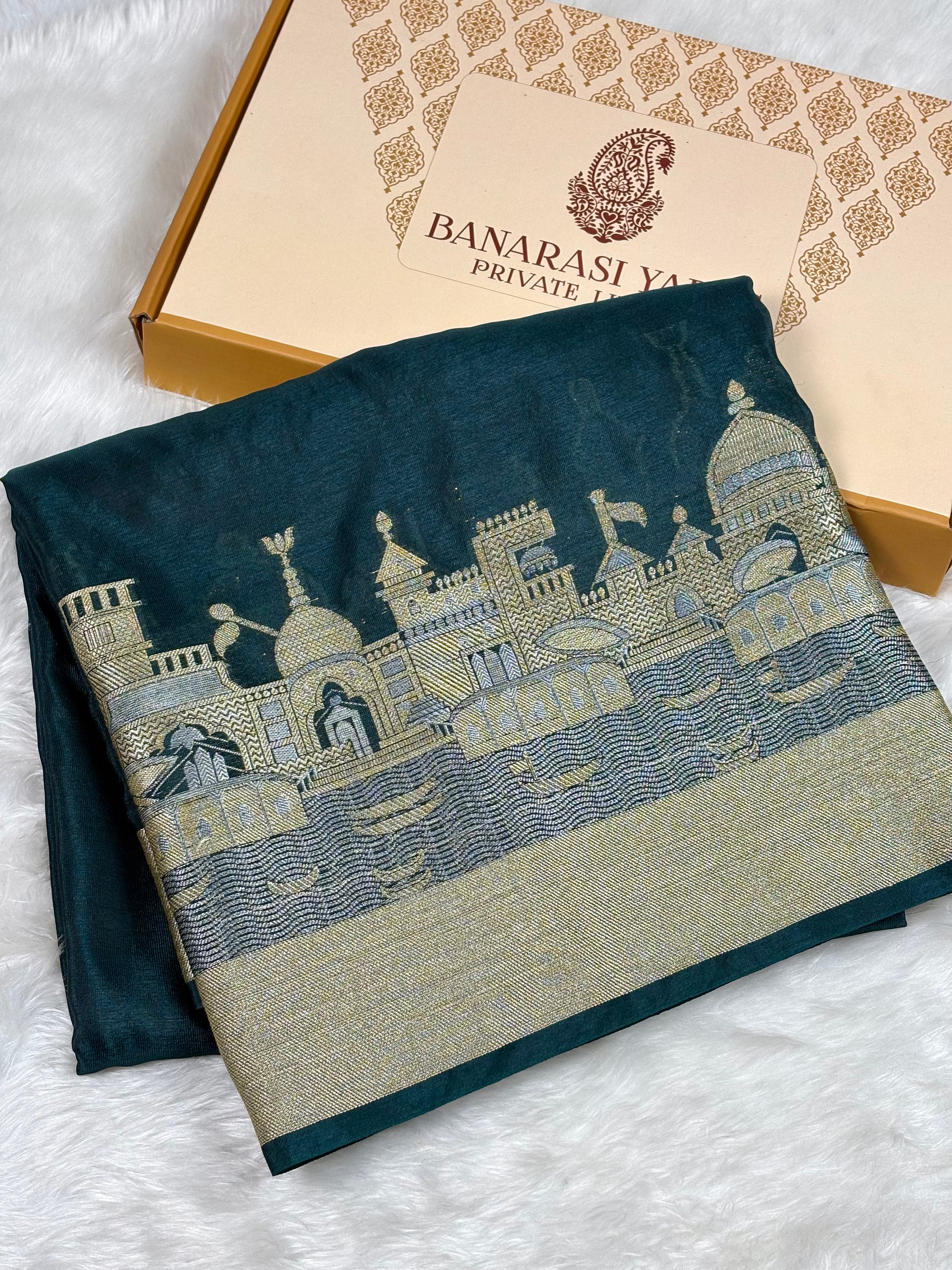 A folded dark teal Banarasi silk saree with a detailed alfi mina zari architectural weaving of a palace, displayed next to its branded packaging box on a white faux fur surface.