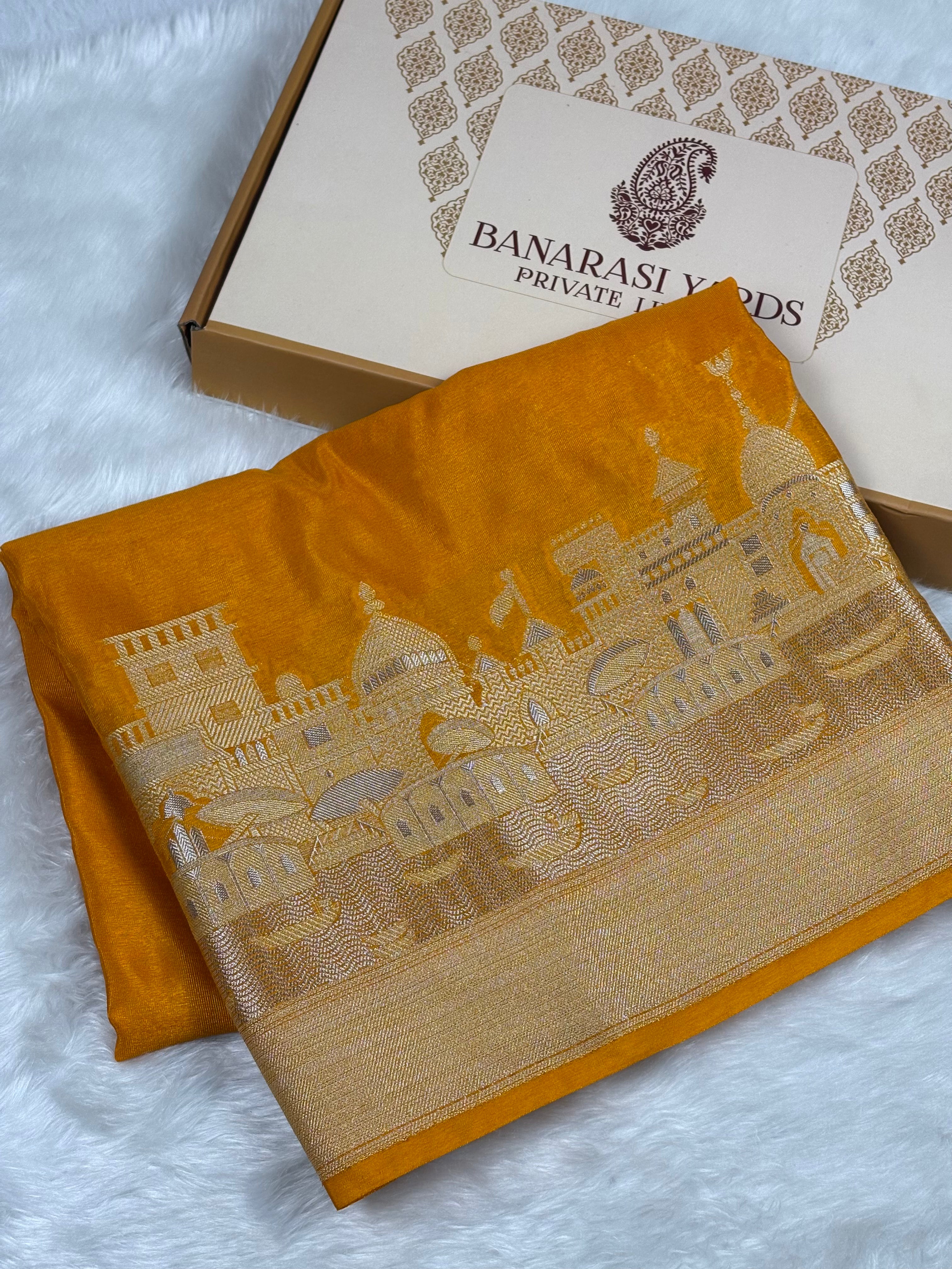 A folded orange silk saree with alifi mina Banarasi City motif, displayed next to its branded 'Banarasi Yards' gift box on a white faux fur background.