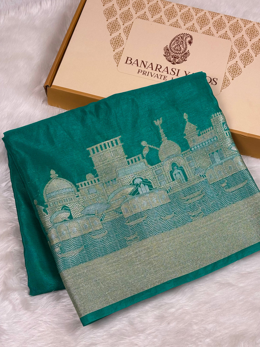 A folded teal green saree with a detailed alfi mina zari architectural pattern, displayed next to its Banarasi Yards Private Limited brand box on a white faux fur background.