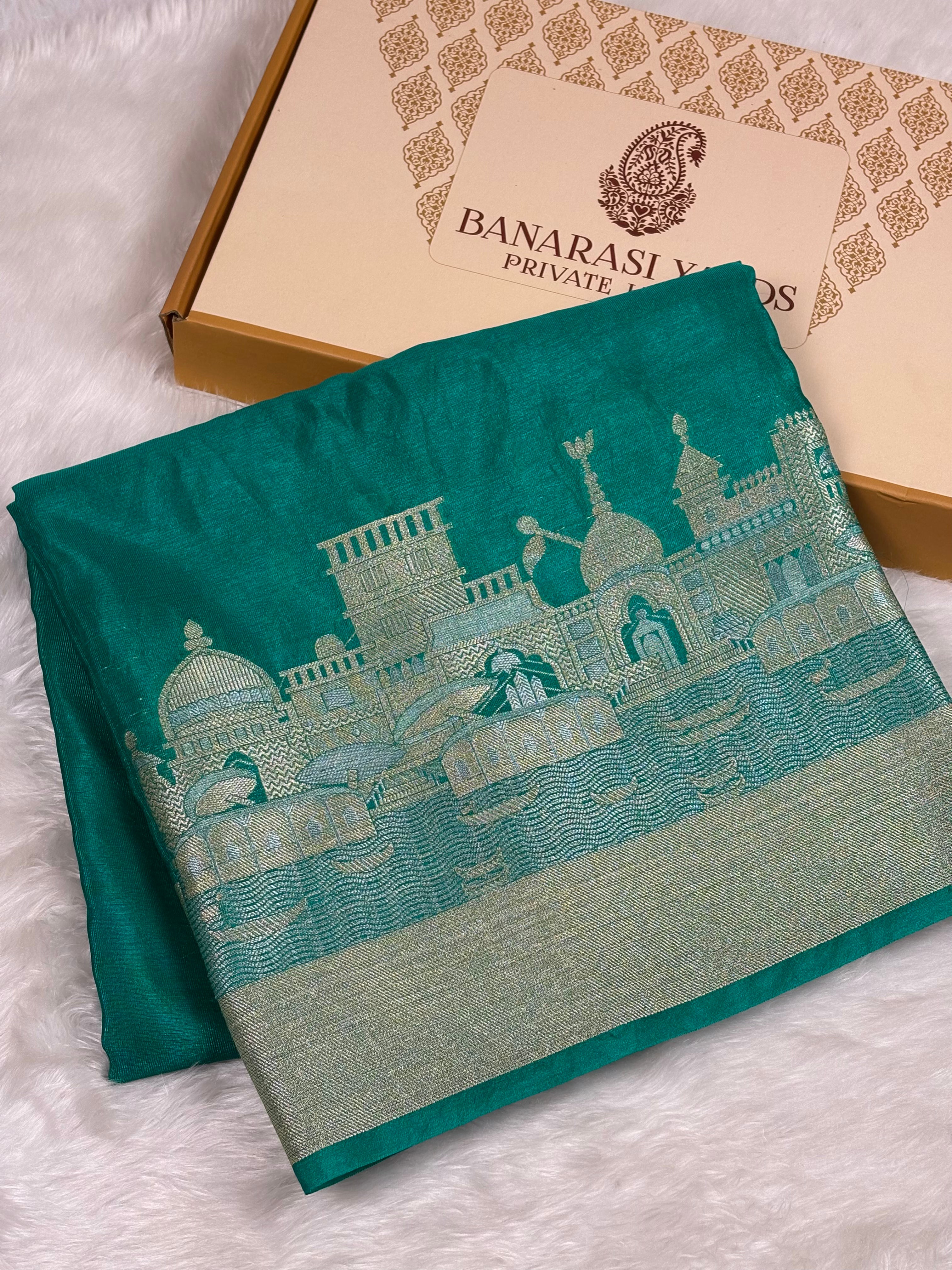 A folded teal green saree with a detailed alfi mina zari architectural pattern, displayed next to its Banarasi Yards Private Limited brand box on a white faux fur background.