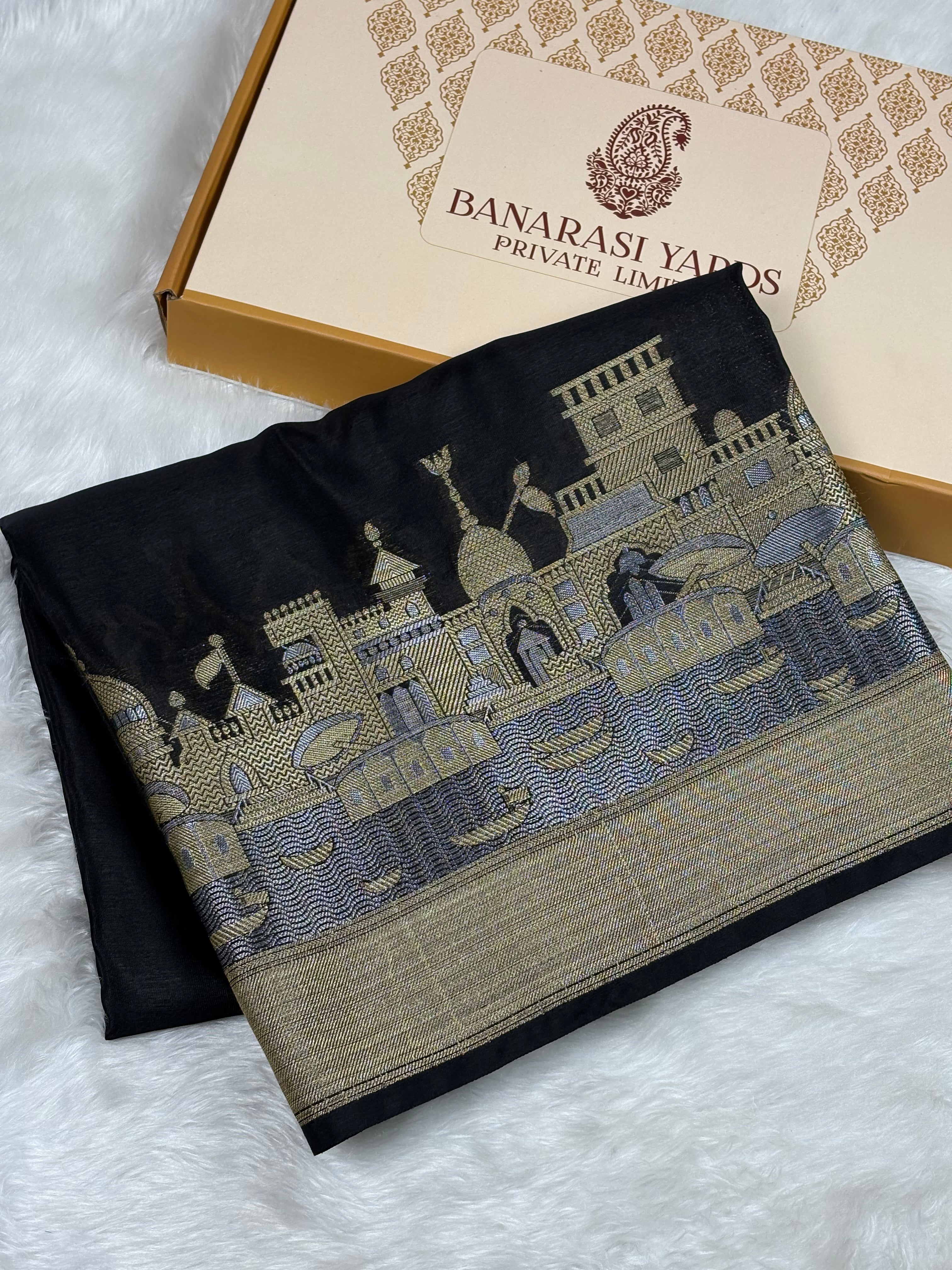 A folded black warm georgette silk saree with a detailed alfi mina zari weaving of a traditional palace and river scene, displayed next to its branded gift box on a white faux fur background.