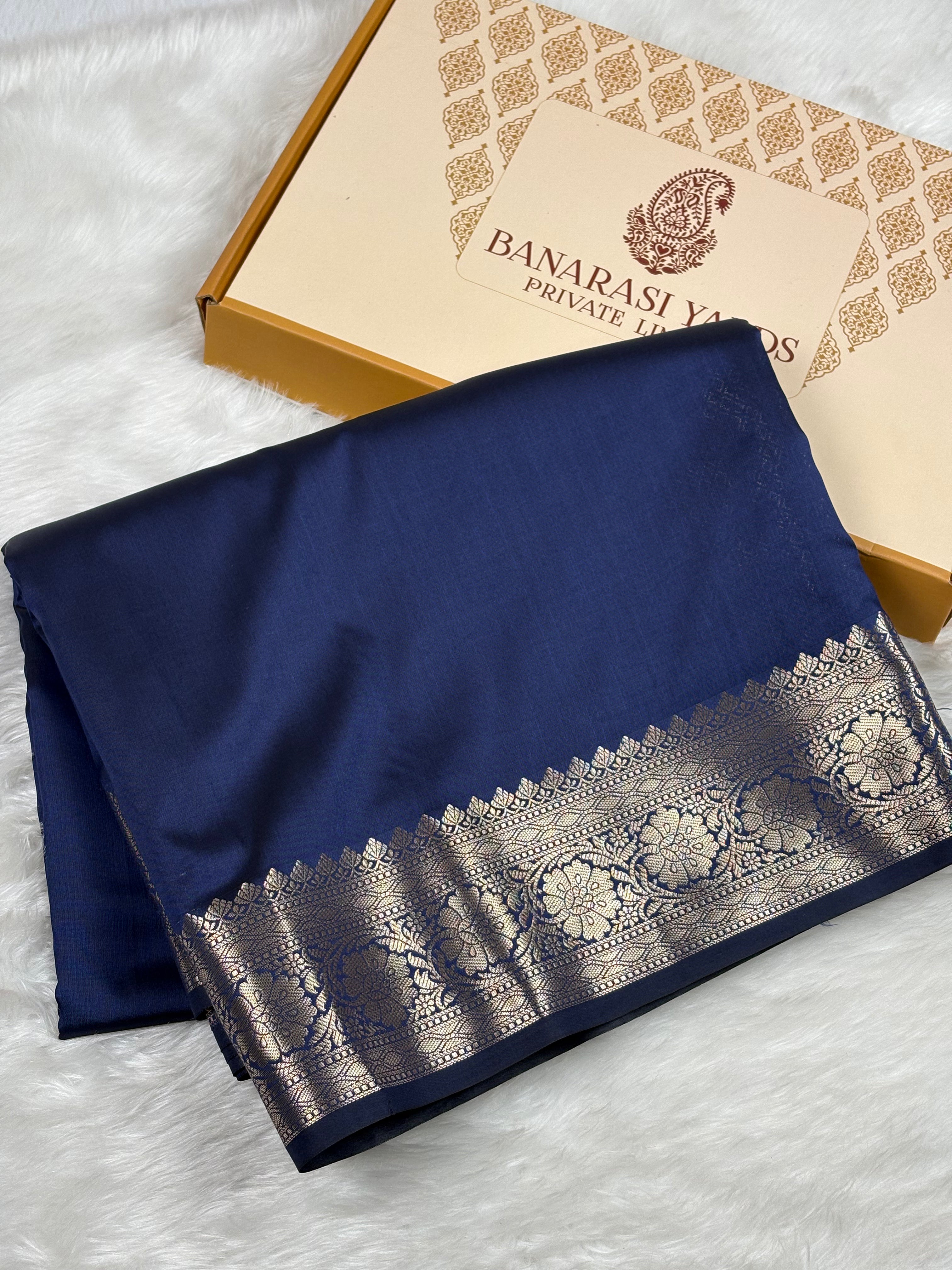 A folded navy blue silk saree with a wide metallic gold border, displayed next to its Banarasi Yards Private Limited box on a white faux fur background.