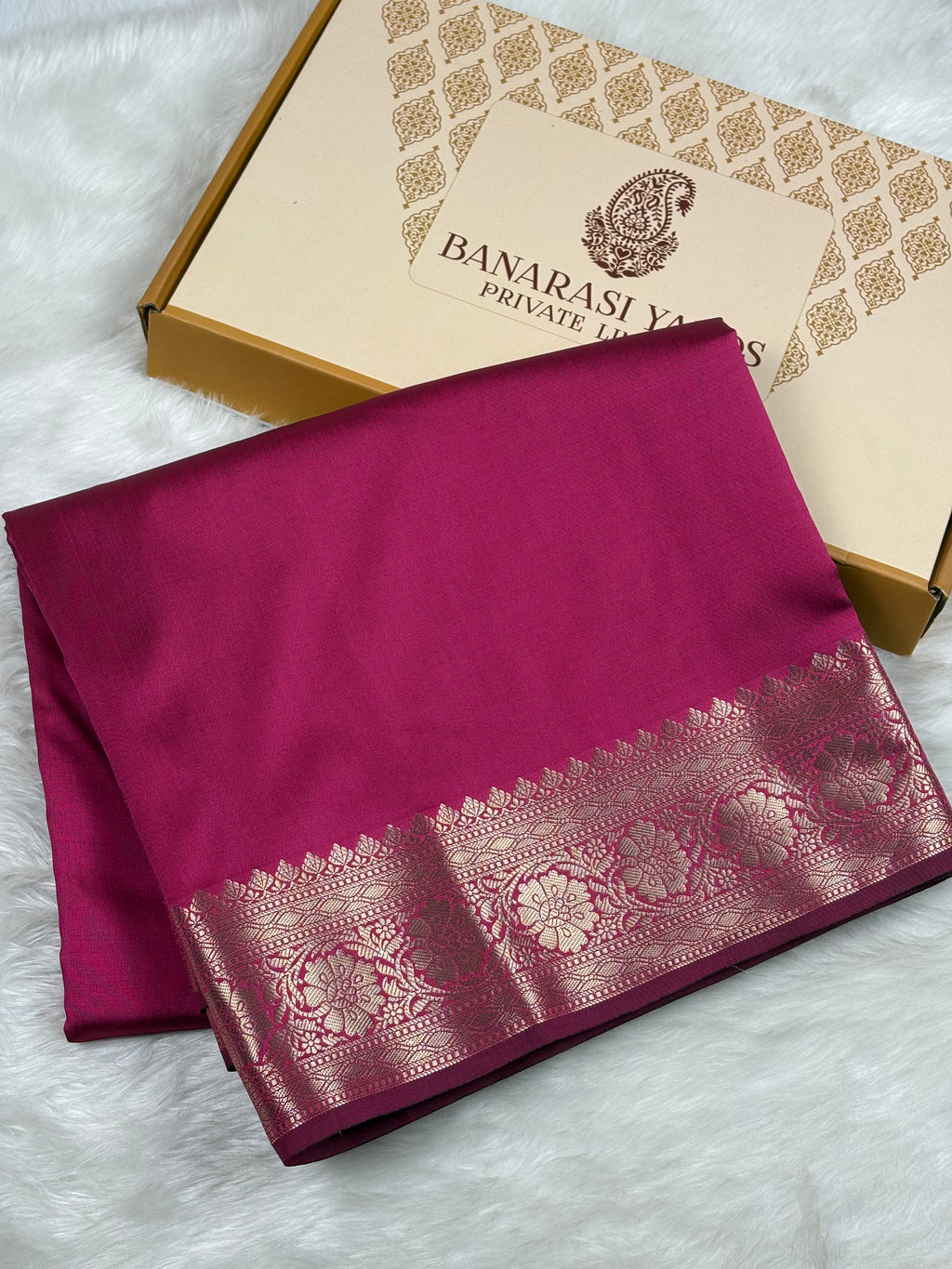A folded magenta silk saree with a wide gold zari border, displayed next to its branded Banarasi Yards Private Limited box on a white faux fur background.