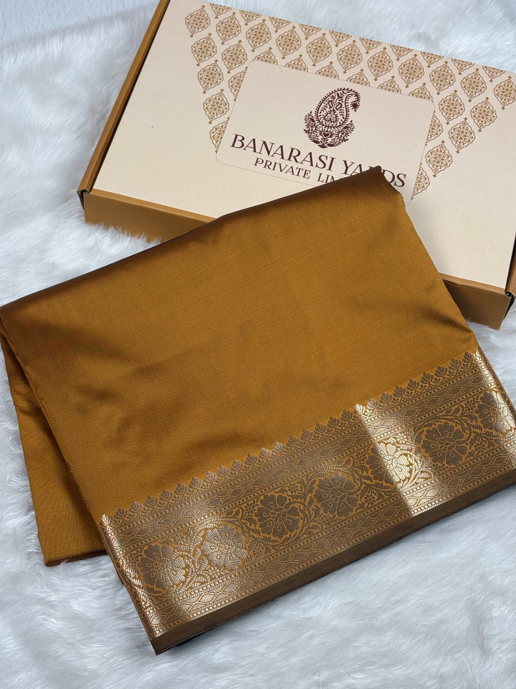 A folded mustard-gold Banarasi silk saree with a detailed metallic gold border, displayed next to its branded packaging box on a white faux fur surface.