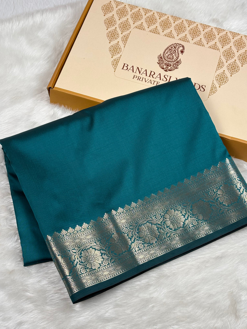 A folded teal silk saree with a wide gold Banarasi border, displayed next to its gift box on a white faux fur background. The box is branded 'BANARASI YARDS PRIVATE LIMITED"