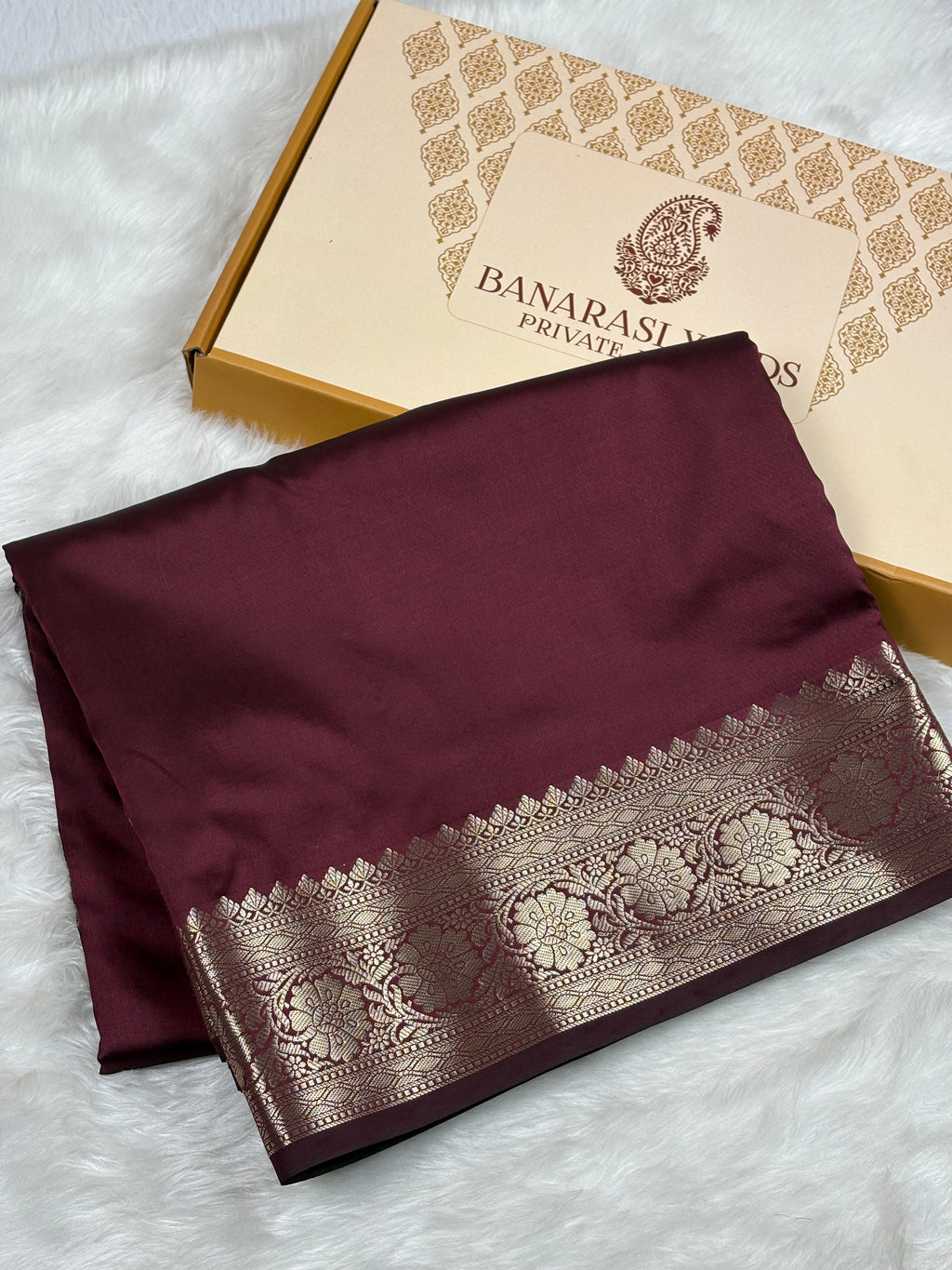 A folded deep maroon silk saree with a wide gold border, displayed next to its branded 'Banarasi Yards Private Limited' gift box on a white faux fur surface.