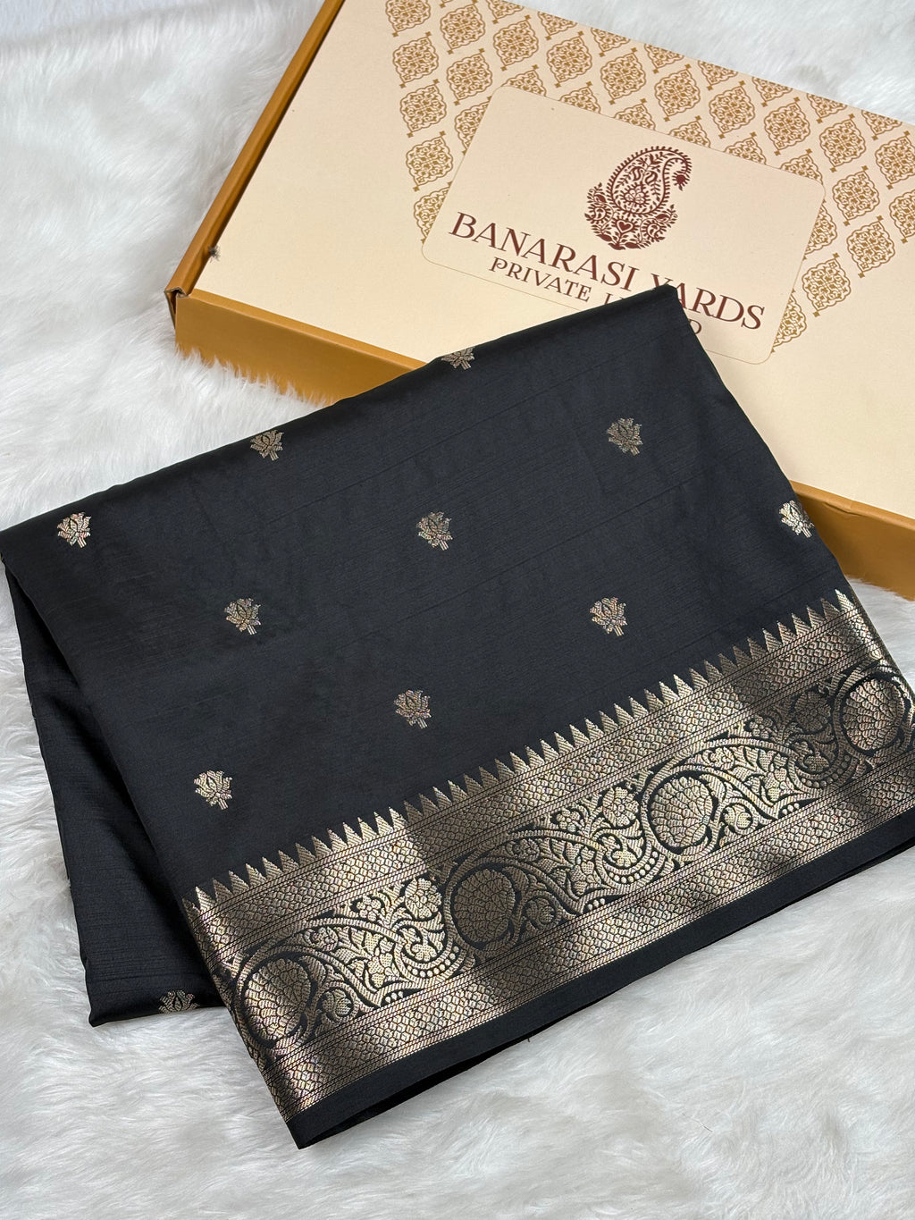 A folded black saree with a wide gold border and small gold motifs, displayed next to its Banarasi Yards Private Limited box on a white faux fur surface.
