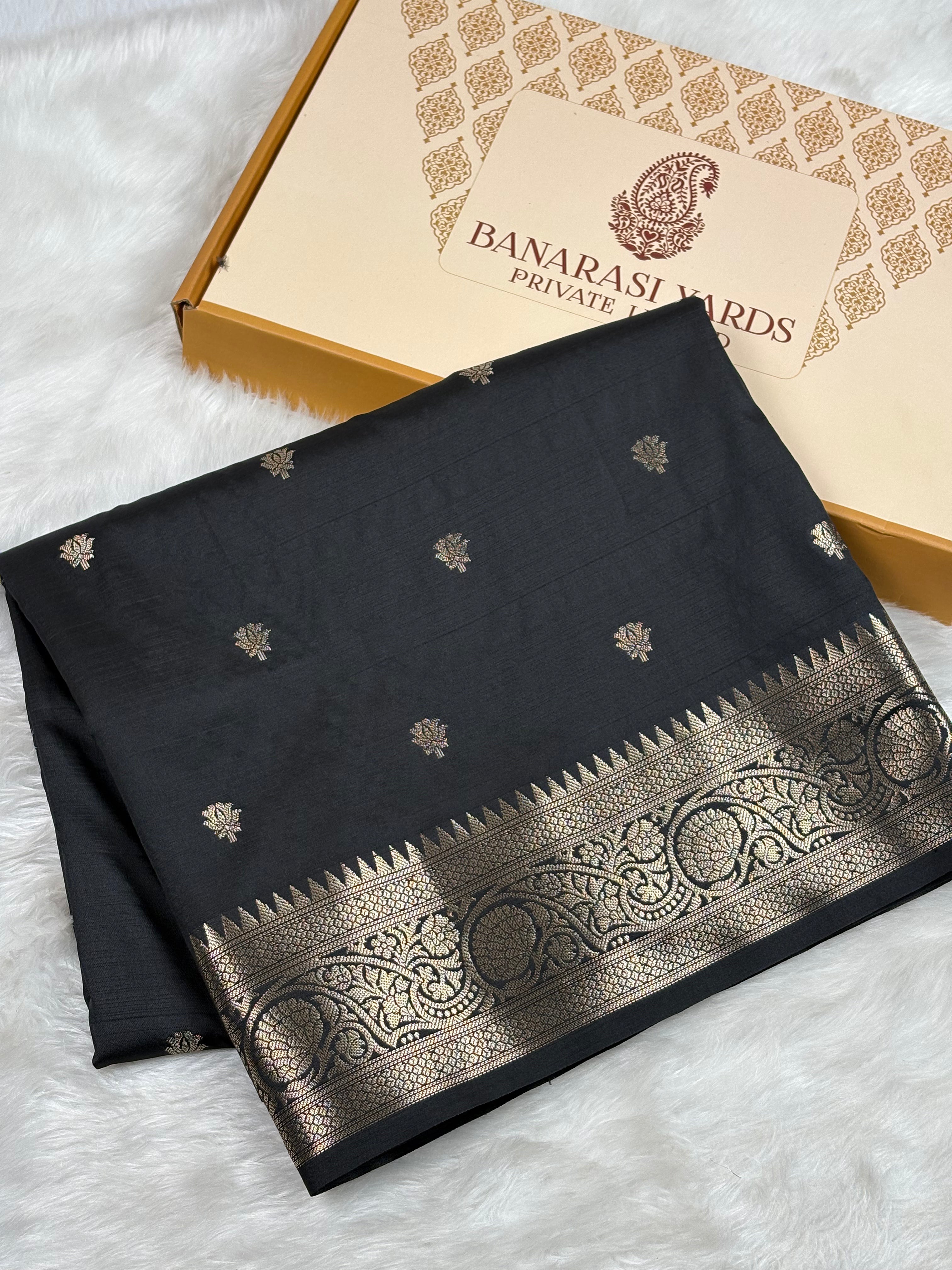 A folded black saree with a wide gold border and small gold motifs, displayed next to its Banarasi Yards Private Limited box on a white faux fur surface.