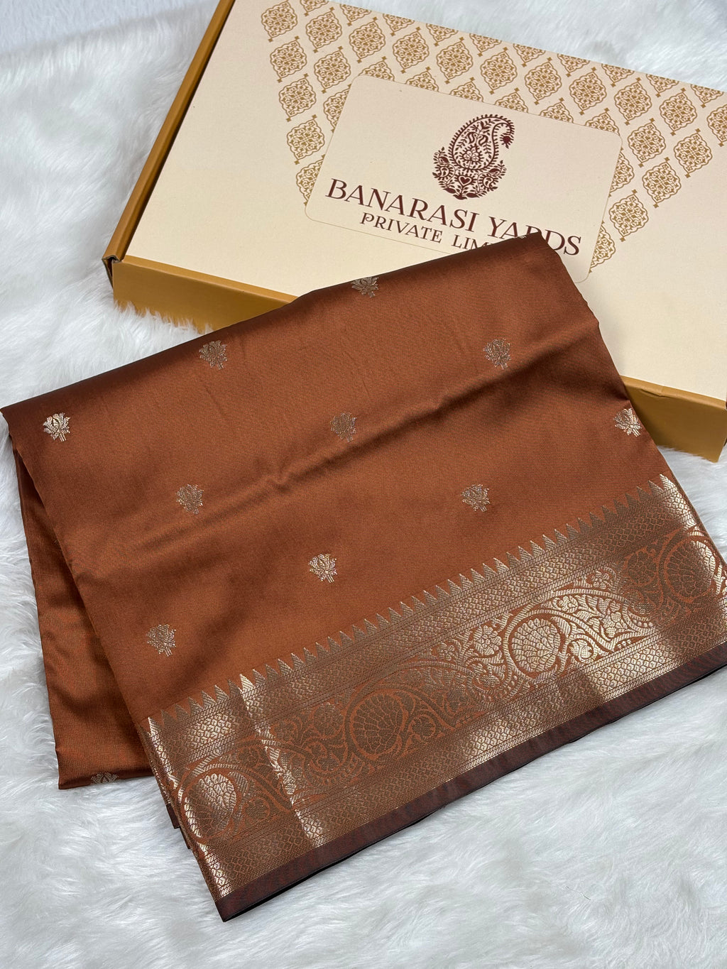 A folded brown Banarasi silk saree with gold zari weaving and a wide ornate border, displayed next to its Banarasi Yards Private Limited gift box on a white faux fur surface.