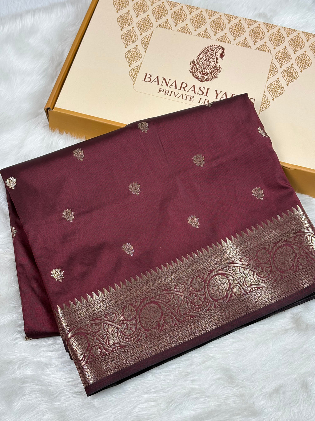 A folded maroon silk saree with gold zari weaving and a wide decorative border, displayed next to its branded packaging box on a white faux fur surface.