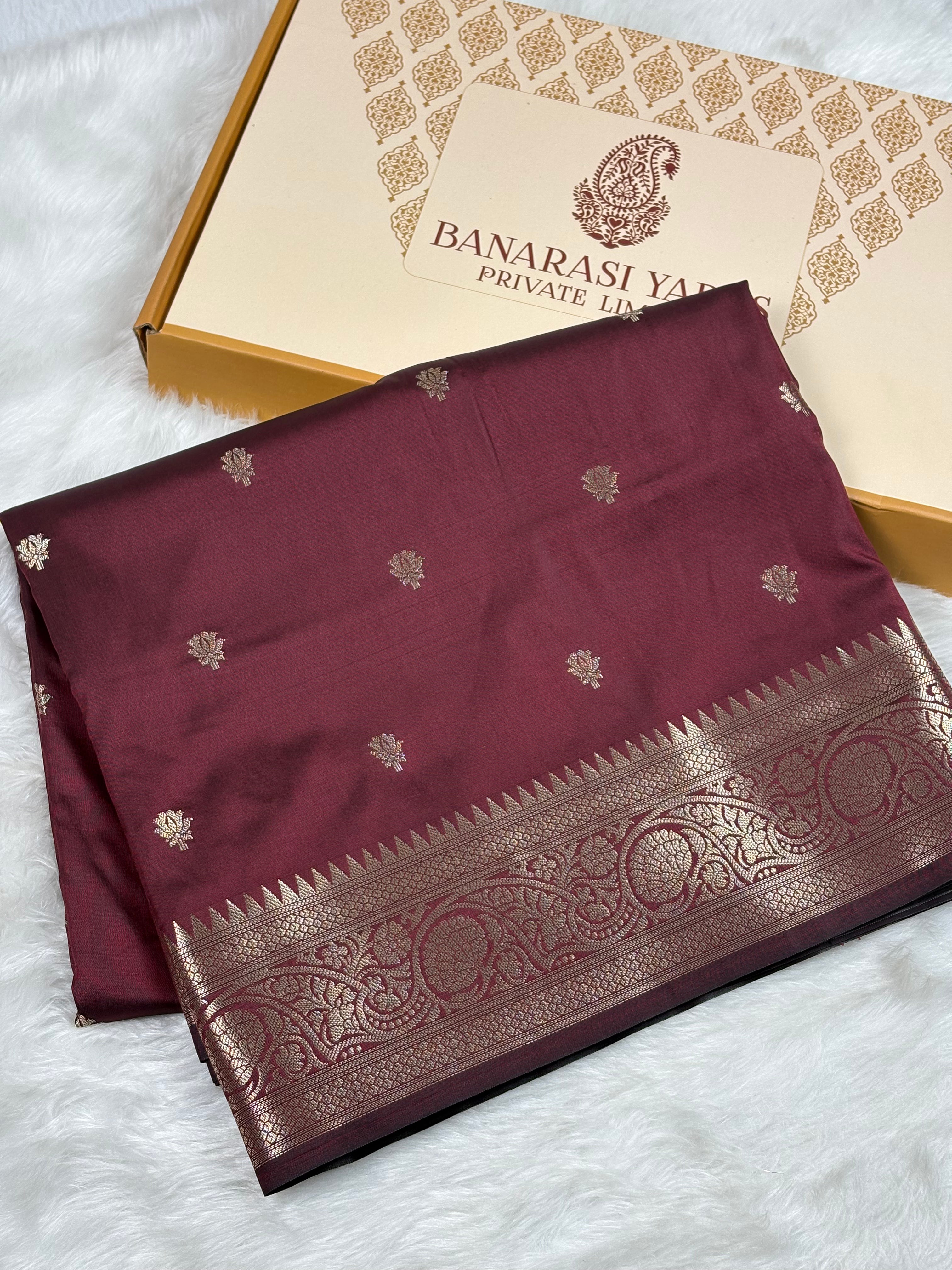 A folded maroon silk saree with gold zari weaving and a wide decorative border, displayed next to its branded packaging box on a white faux fur surface.