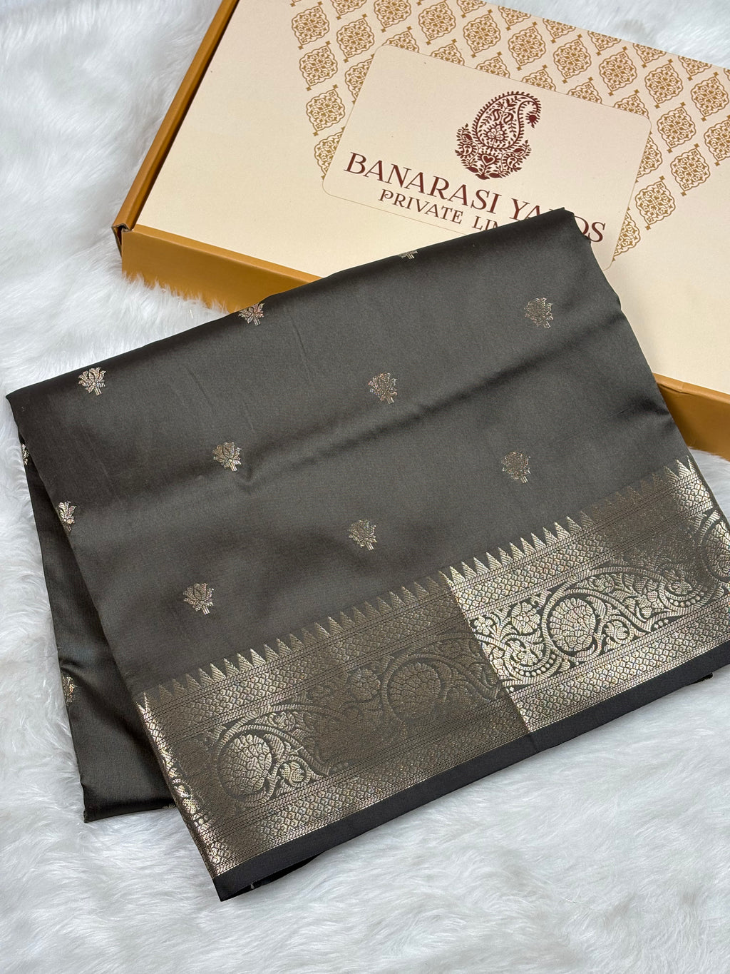 A folded dark gray Banarasi silk saree with gold zari weaving and a wide ornate gold border, displayed next to its Banarasi Yards Private Limited gift box on a white faux fur background.
