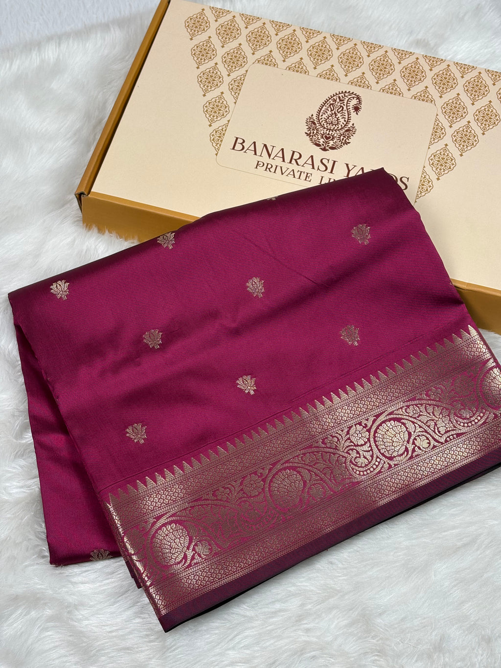 A folded deep magenta silk saree with gold zari motifs and a wide gold border, displayed next to its branded Banarasi Yards Private Limited box on a white faux fur surface.