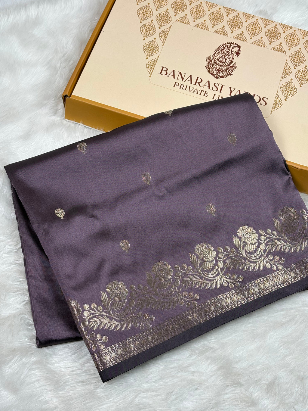 A folded dark purple Banarasi silk saree with gold floral patterns and borders, displayed next to its Banarasi Yards Private Limited box on a white faux fur background.