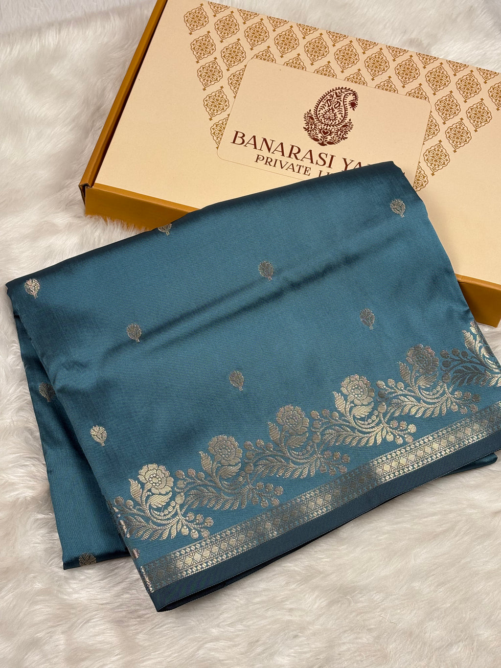 A folded teal blue Banarasi silk saree with gold zari floral patterns and borders, displayed next to its branded gift box on a white faux fur surface.