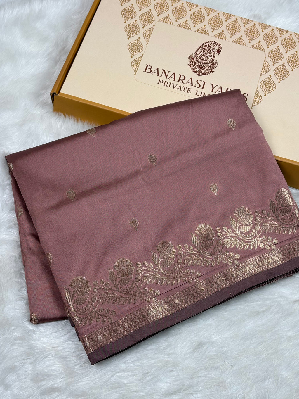 A folded dusty rose silk saree with gold zari floral border and motifs, displayed next to its Banarasi Yards Private Limited gift box on a white faux fur background.