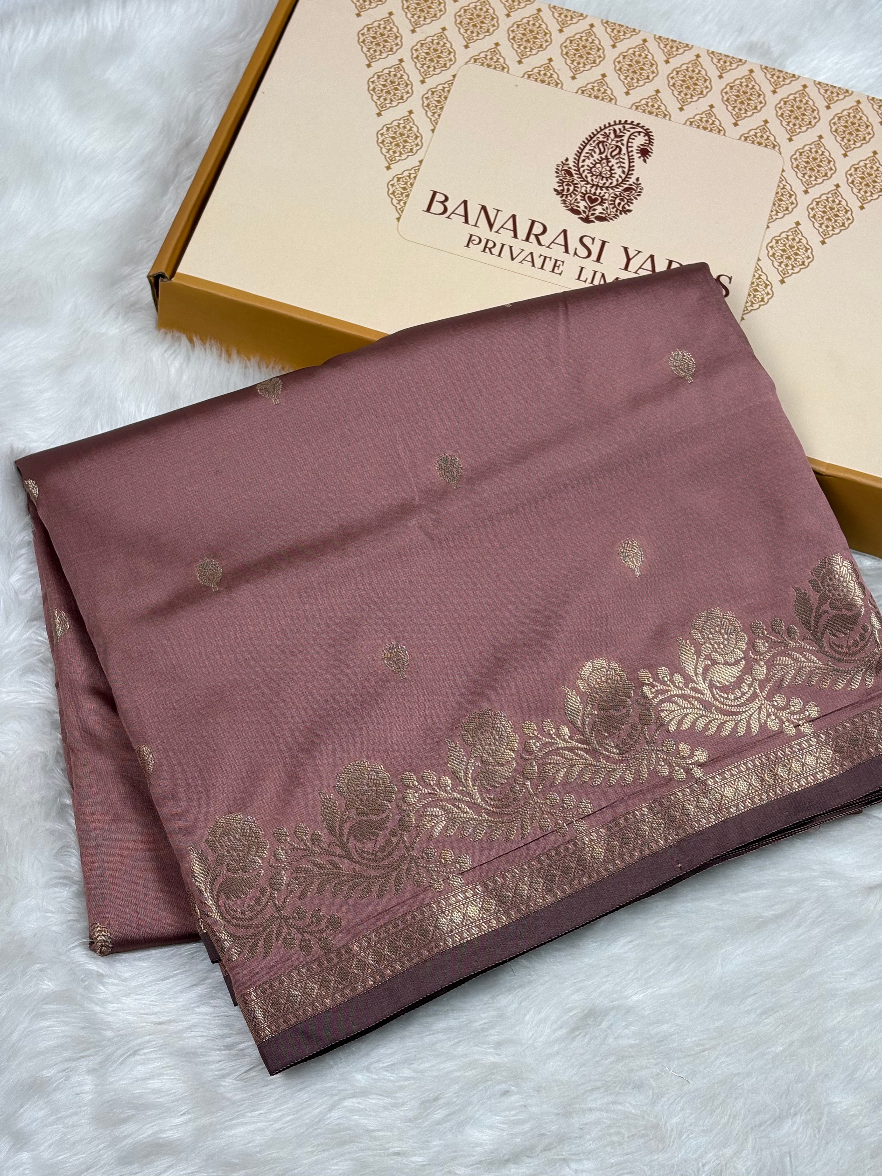 A folded dusty rose silk saree with gold zari floral border and motifs, displayed next to its Banarasi Yards Private Limited gift box on a white faux fur background.