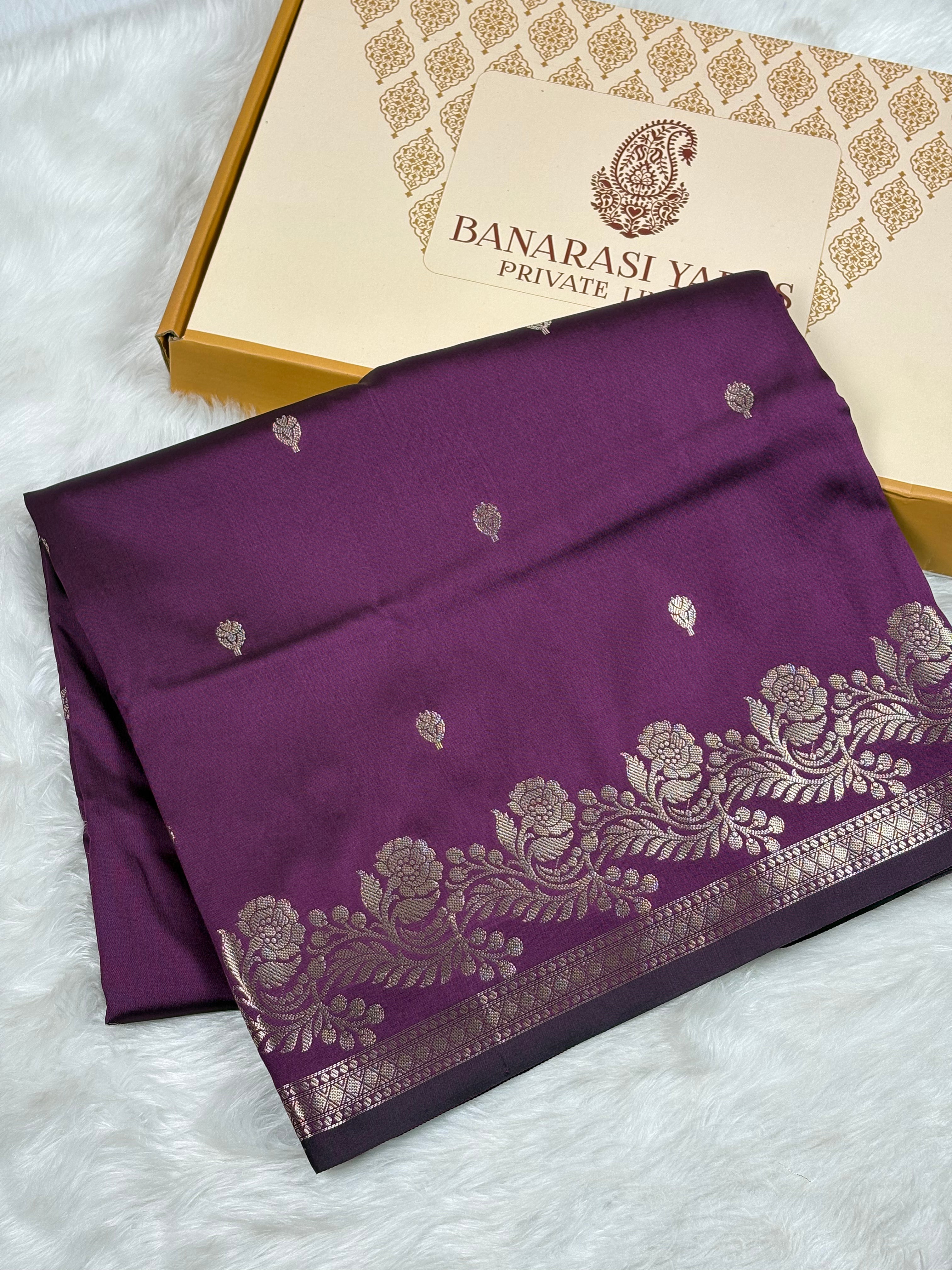 A folded deep purple Banarasi silk saree with gold floral zari work and border detailing, displayed next to its Banarasi Yards Private Limited gift box on a white faux fur background.
