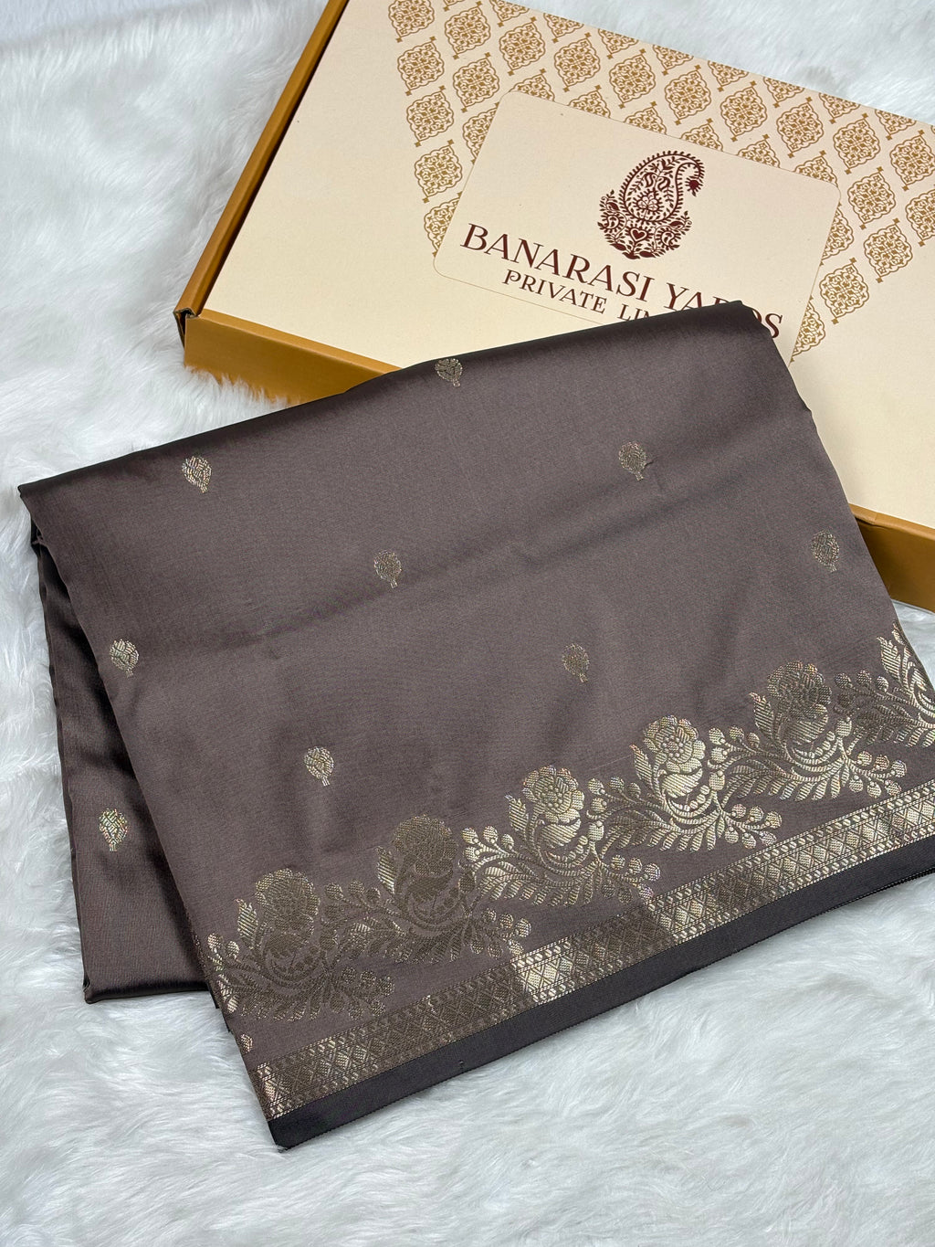 A folded dark brown Banarasi silk saree with gold floral patterns and a border, displayed next to its branded packaging box on a white faux fur surface.