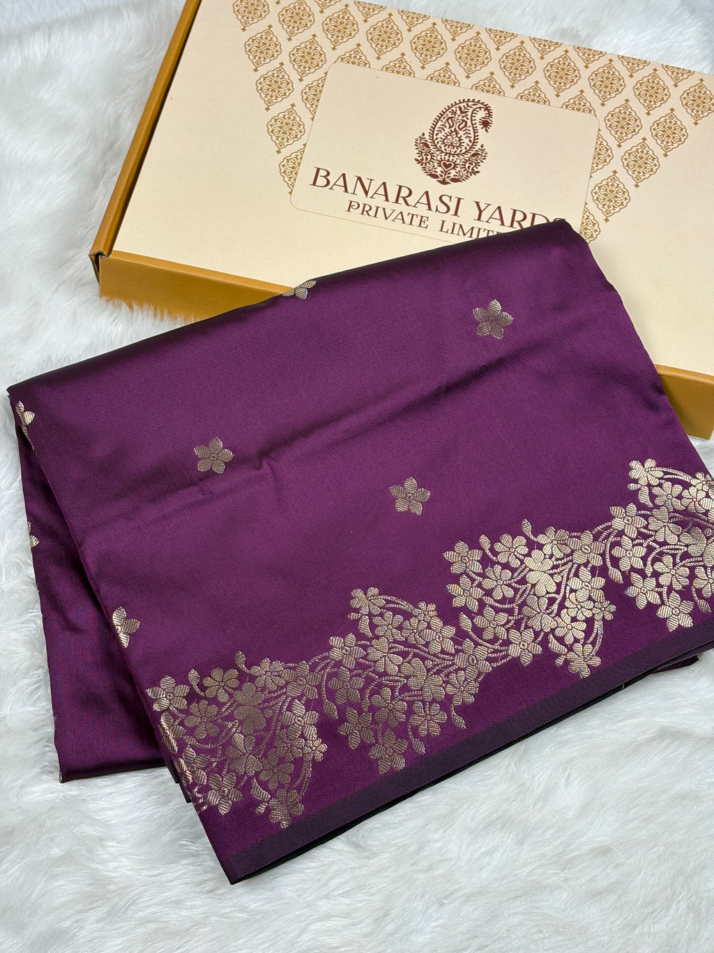 A folded deep purple Banarasi silk saree with gold floral borders and motifs, displayed next to its Banarasi Yards Private Limited box on a white faux fur background.