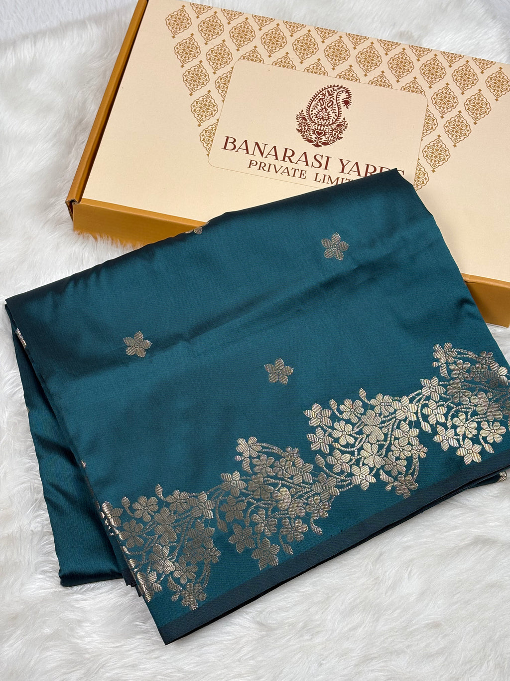 A folded dark teal blue saree with gold floral zari weaving, displayed next to its Banarasi Yards Private Limited box on a white faux fur background.