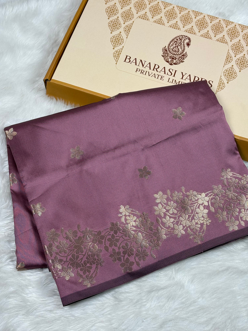 A folded purple Banarasi silk saree with gold floral patterns and a border, displayed next to its branded packaging box on a white faux fur surface.