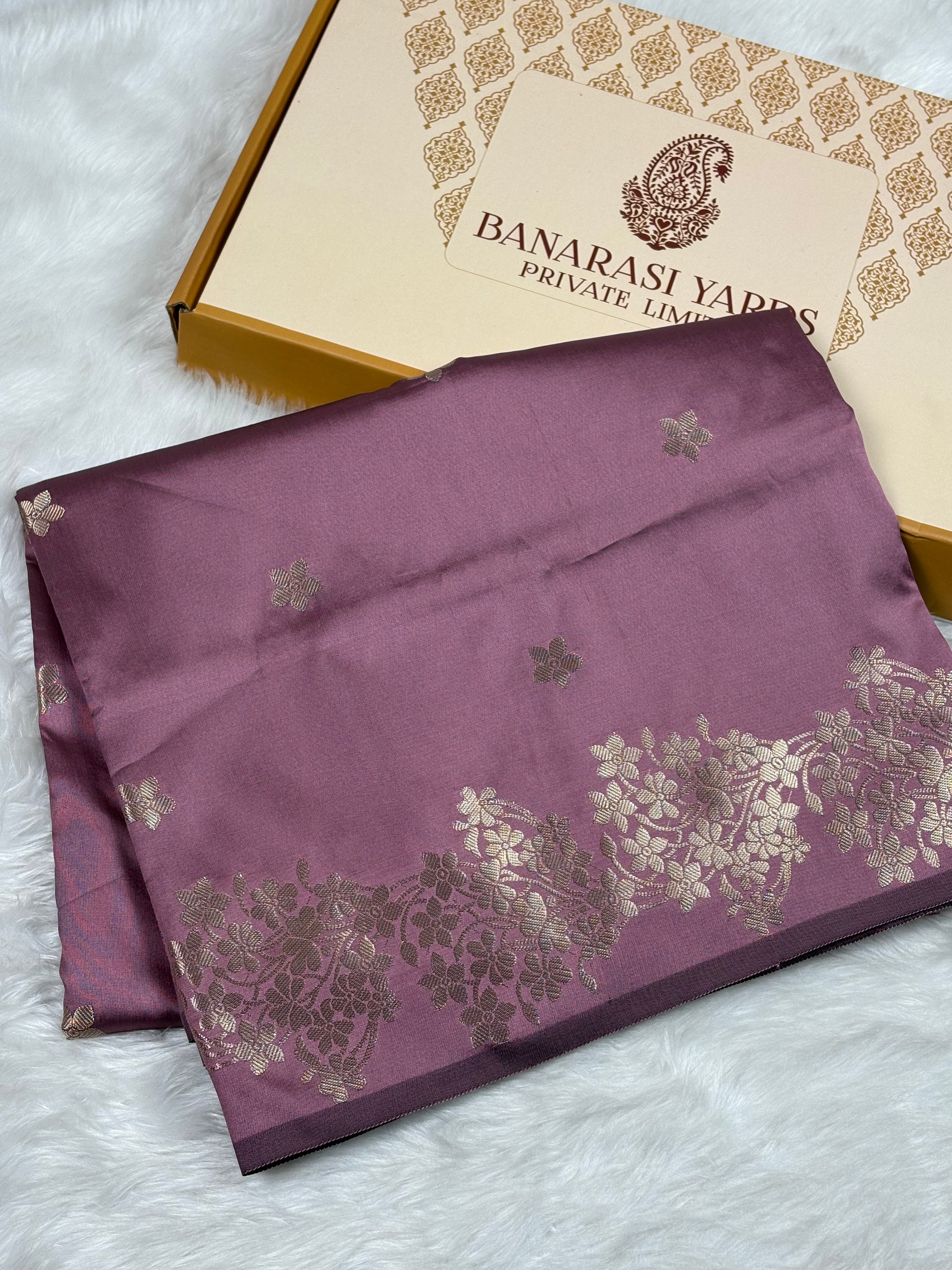 A folded purple Banarasi silk saree with gold floral patterns and a border, displayed next to its branded packaging box on a white faux fur surface.