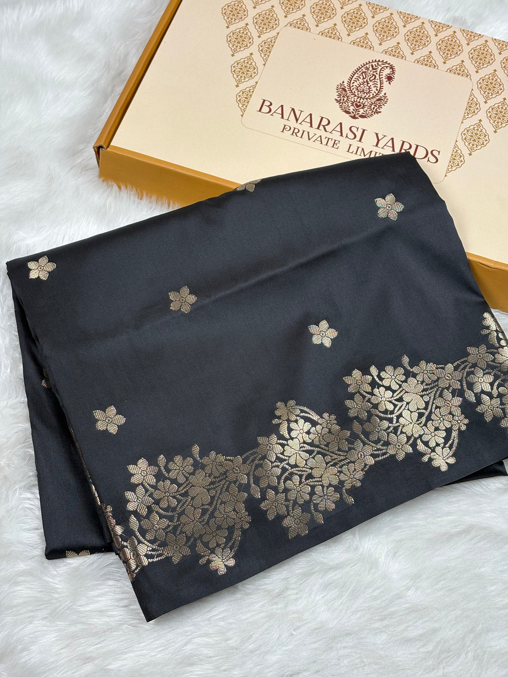 A folded black silk saree with gold floral patterns and border motifs, displayed next to its Banarasi Yards box on a white faux fur surface.