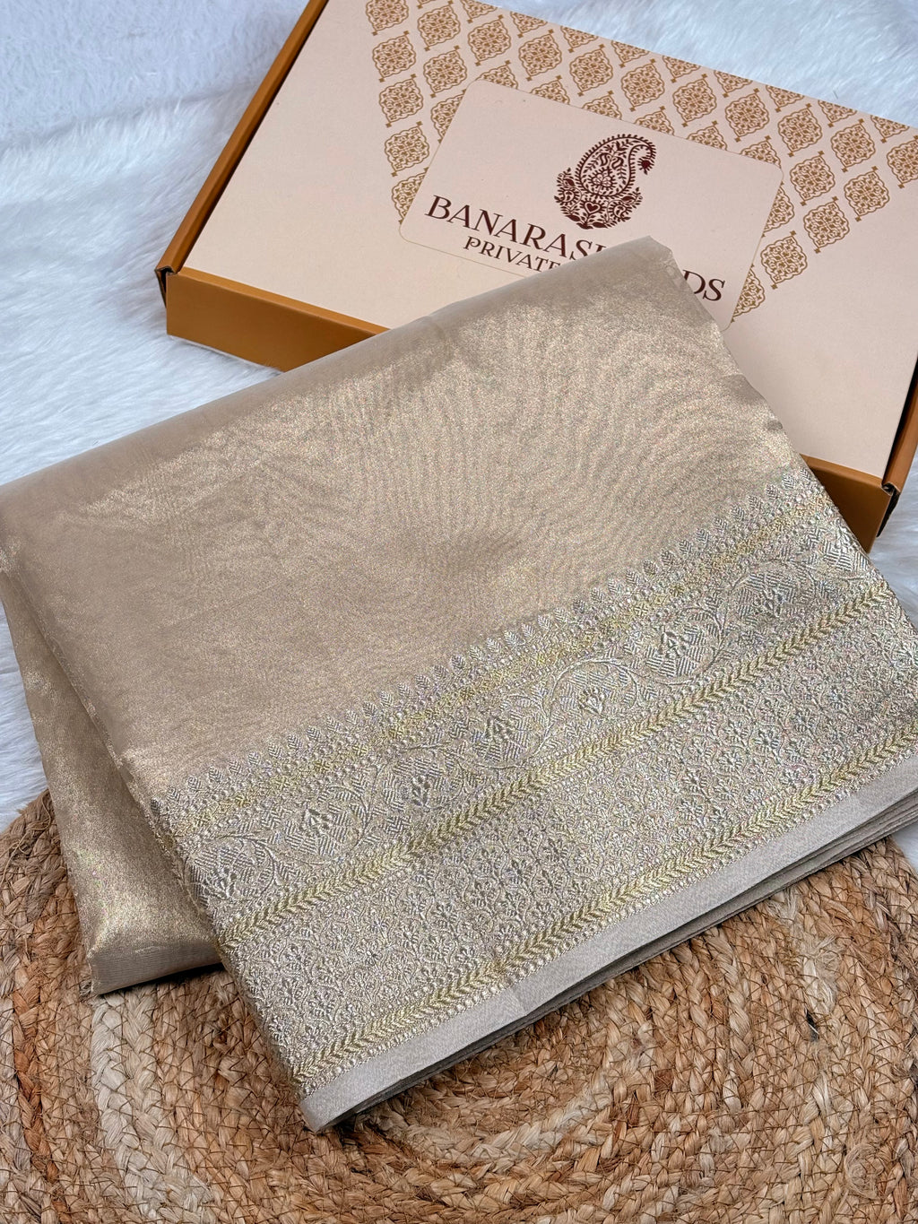 A folded tishu silk saree with sona rupa zari border, displayed on a woven jute mat next to its branded packaging box.