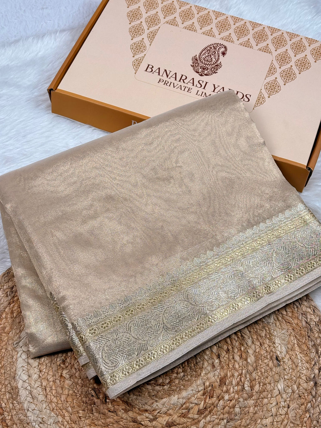 A folded beige Banarasi tishu silk saree with sona rupa weaving border, displayed on a woven jute mat next to its branded packaging box.