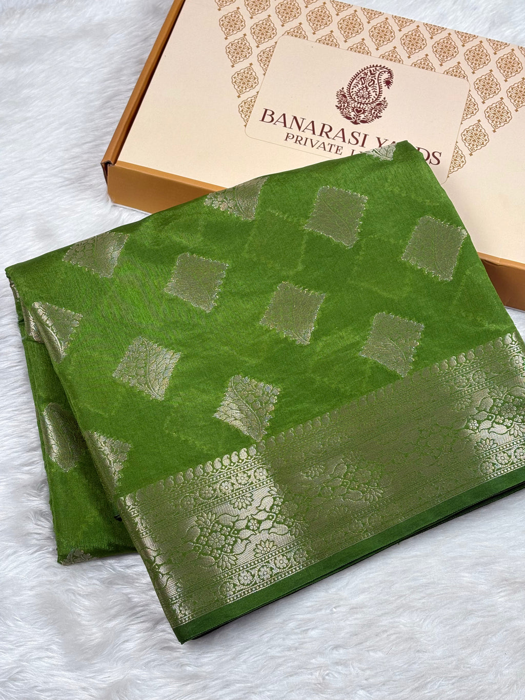 A folded green Banarasi silk saree with gold zari woven patterns and a wide gold border, displayed next to its Banarasi Yards Private Limited box on a white faux fur background.
