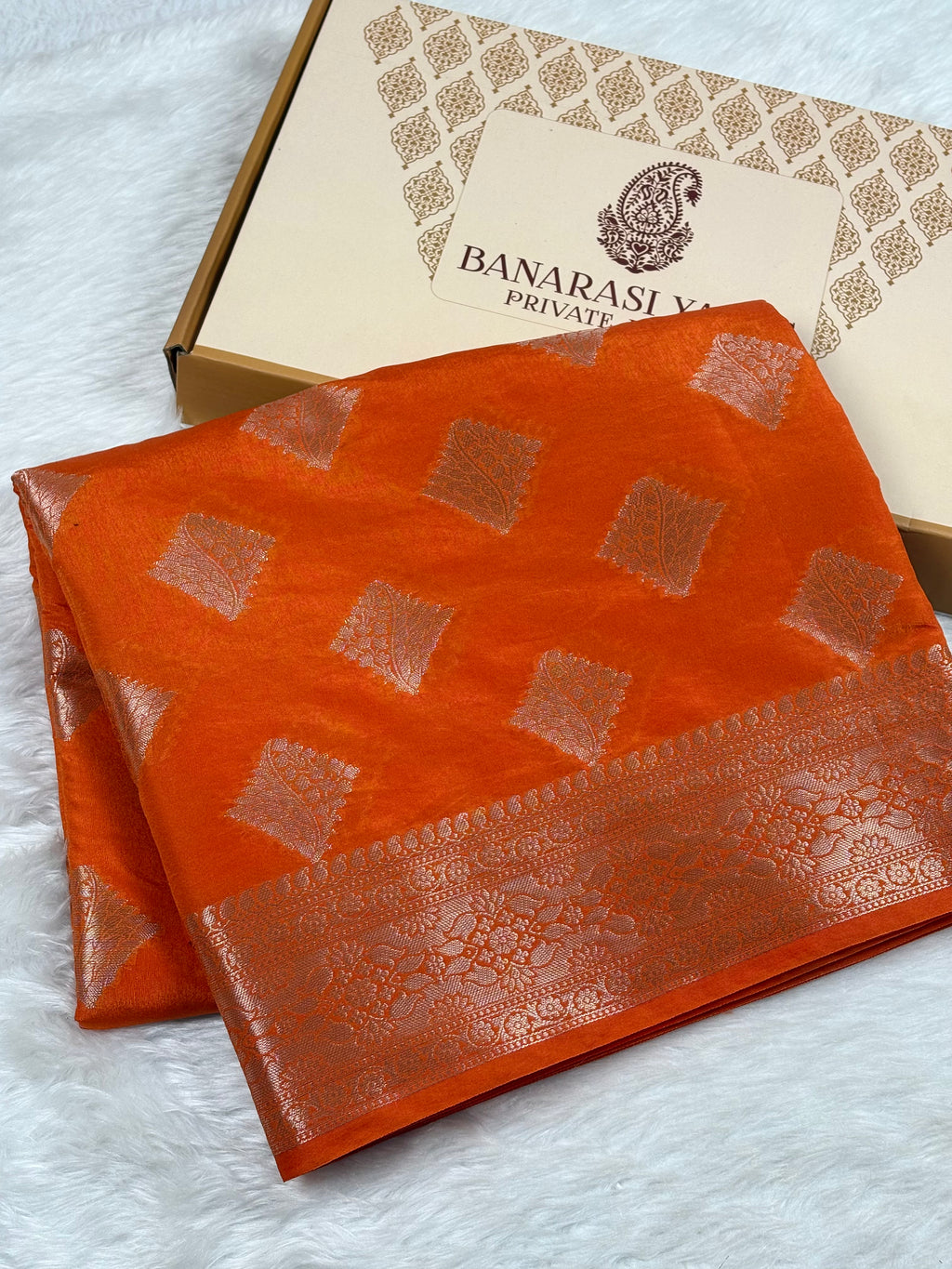An orange Banarasi silk saree with gold woven motifs and a wide gold border, displayed next to its branded packaging box on a white faux fur surface.