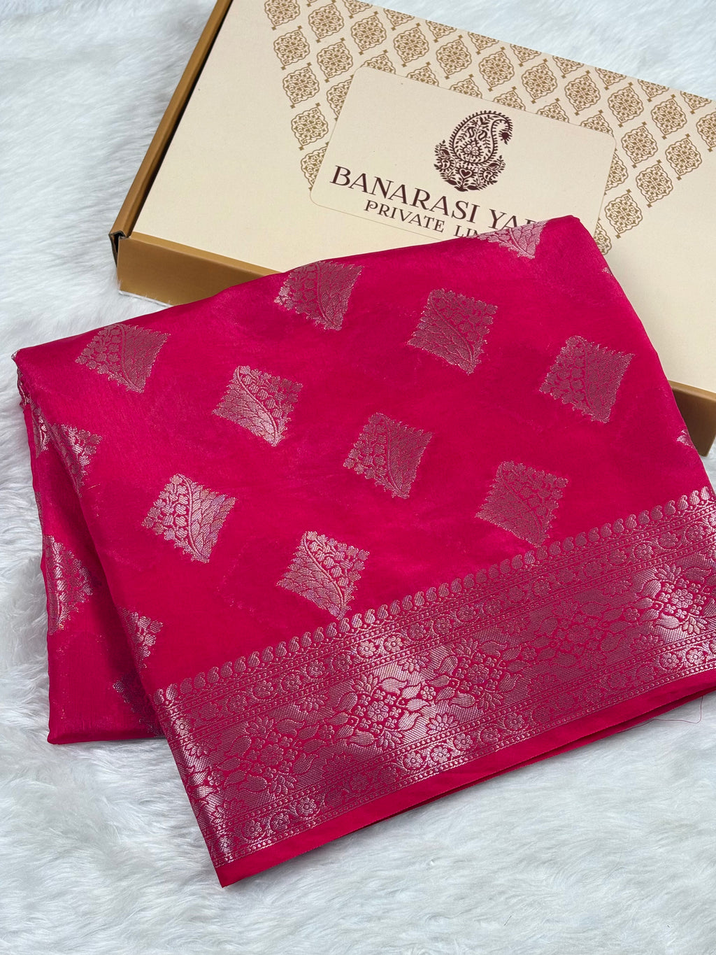 A folded bright pink Banarasi silk saree with gold woven motifs and a border, displayed next to its branded packaging box on a white faux fur surface.