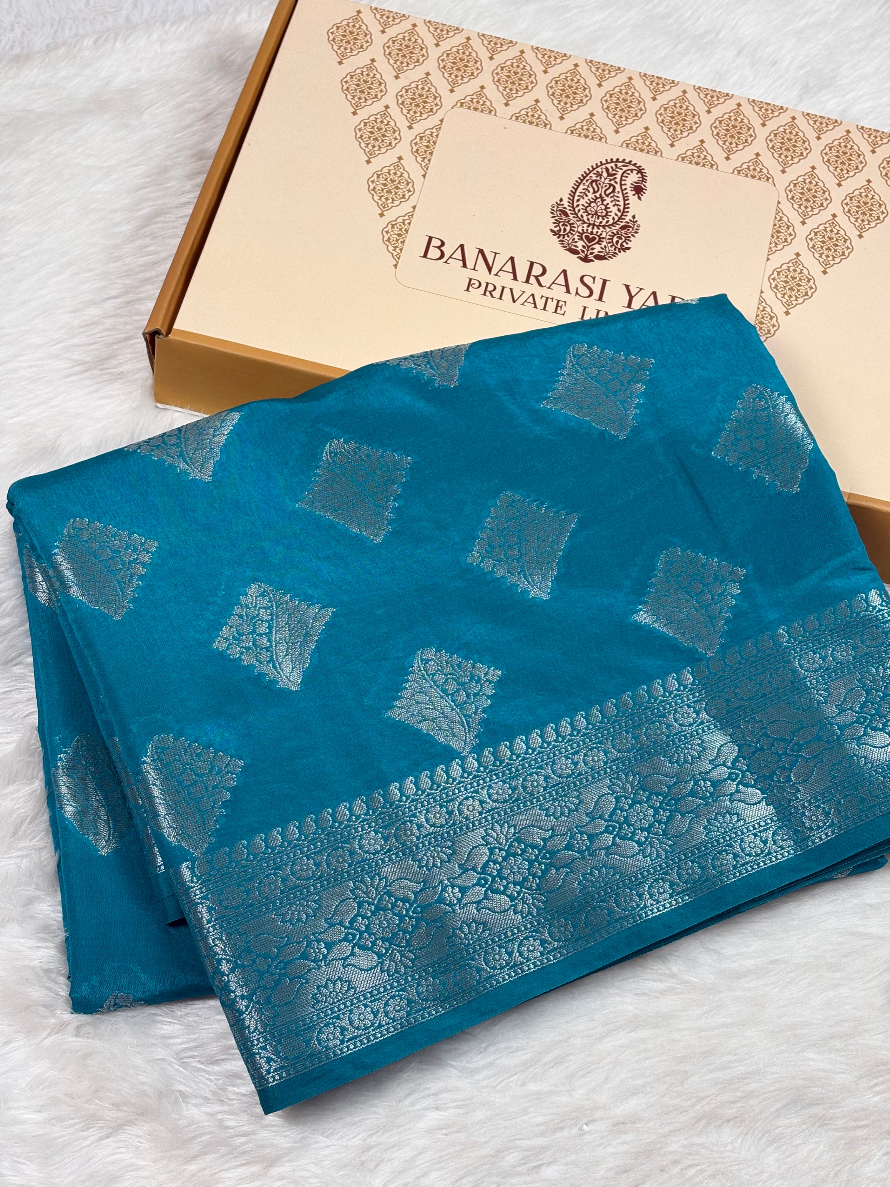 A folded teal blue Banarasi silk saree with silver zari woven geometric patterns and a wide decorative border, displayed next to its branded gift box on a white faux fur surface.