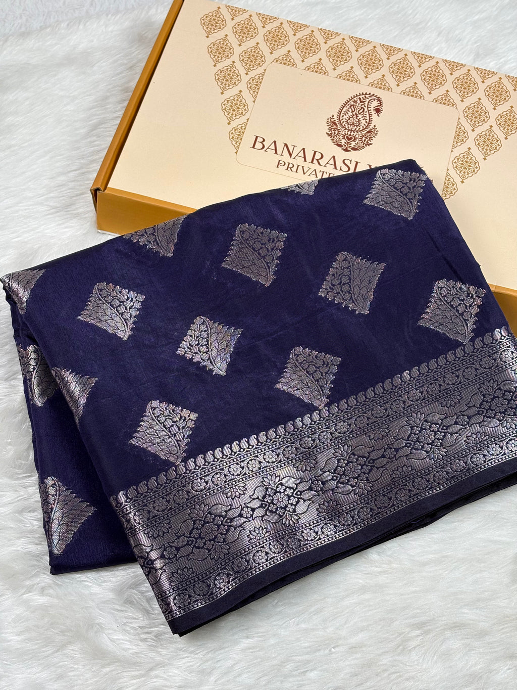 A folded navy blue Banarasi silk saree with silver zari woven geometric patterns and a wide decorative border, displayed next to its Banarasi Yards Private Limited gift box on a white faux fur surface.