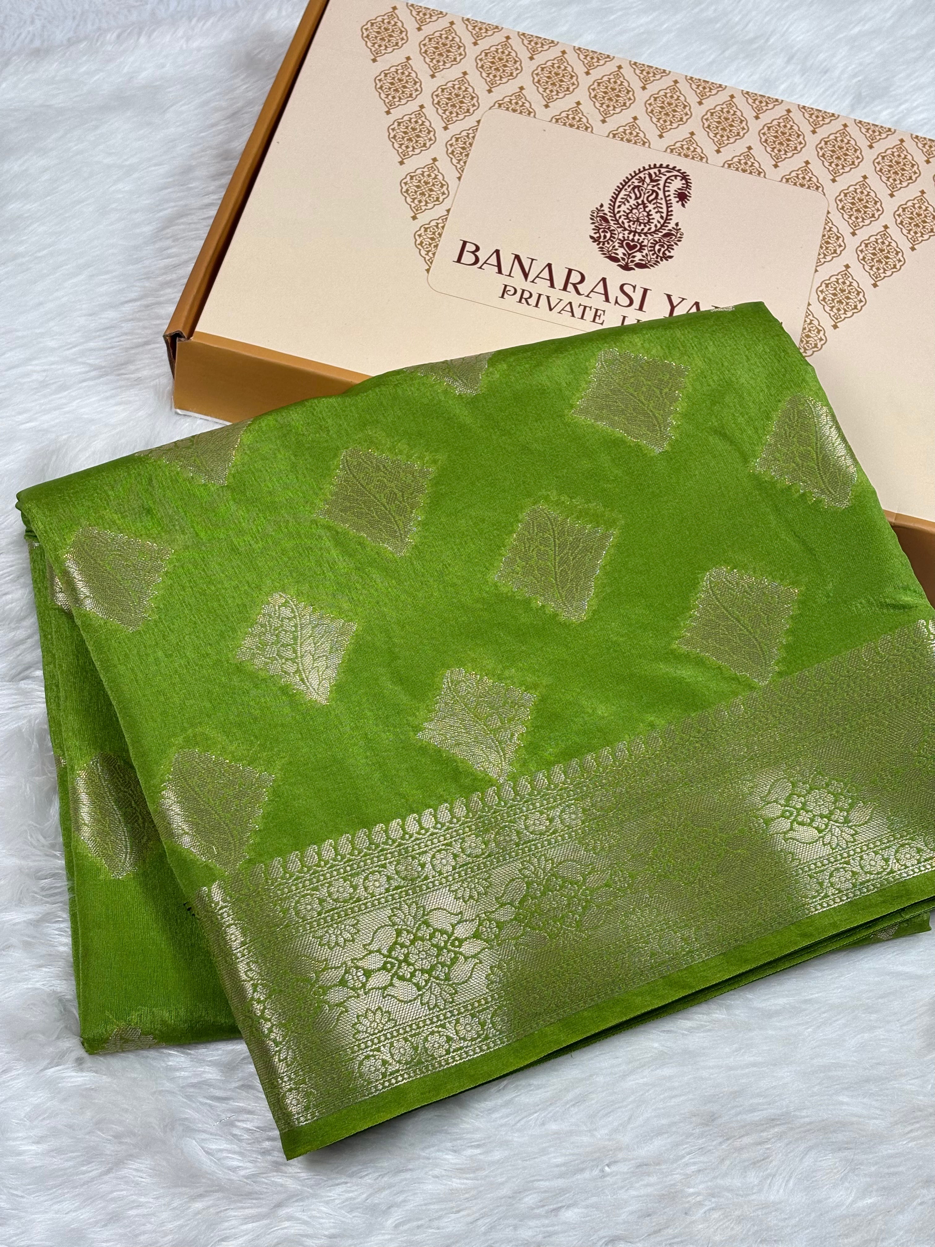 A folded green Banarasi silk saree with gold zari woven geometric patterns and a wide decorative border, displayed next to its branded packaging box on a white faux fur surface.