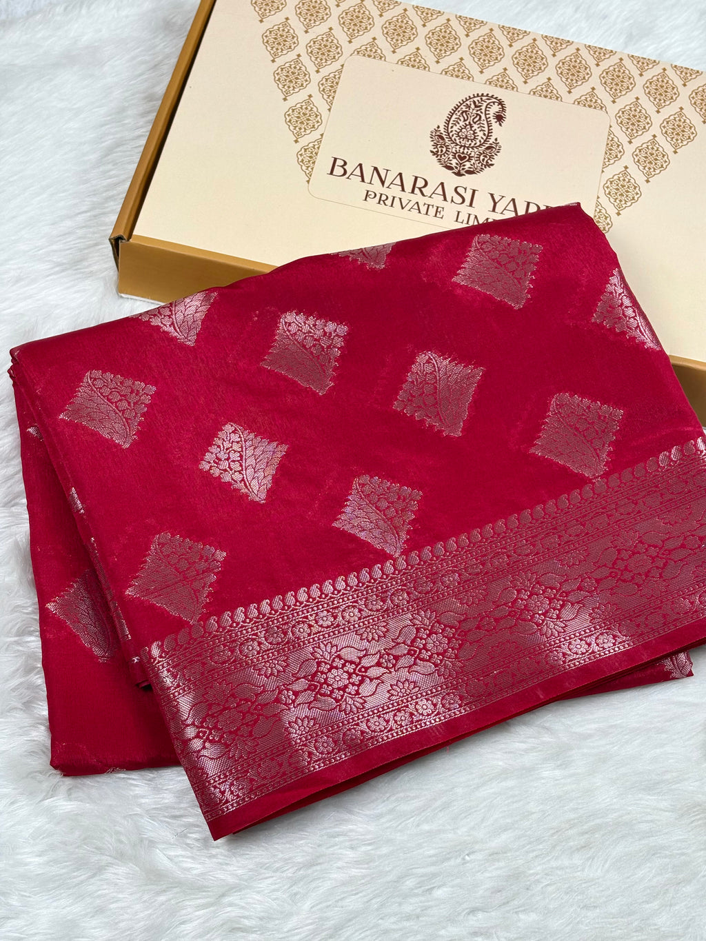 A folded deep red Banarasi silk saree with gold zari woven geometric patterns and a wide decorative border, displayed next to its branded packaging box on a white faux fur surface.