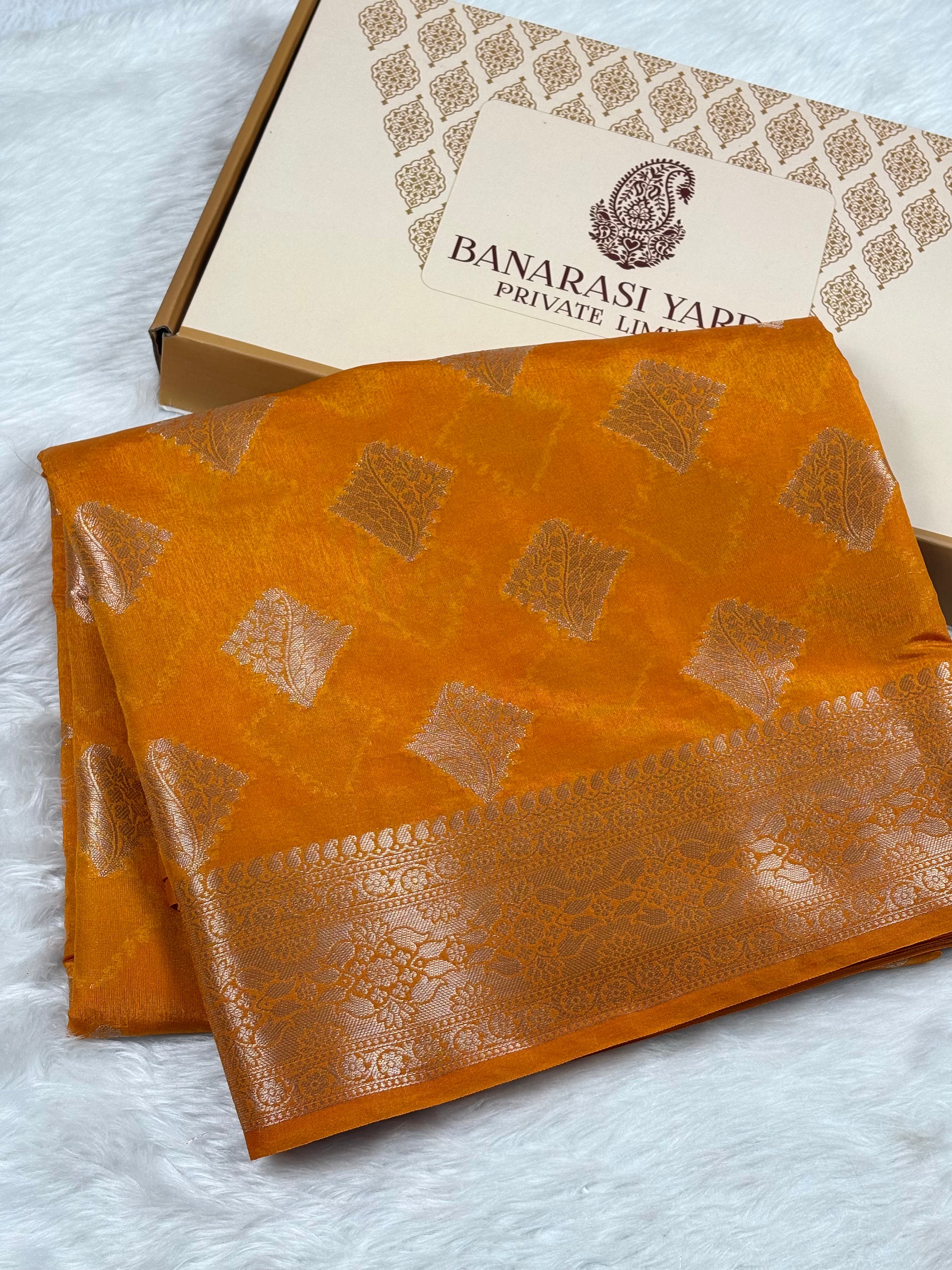 An orange silk saree with gold woven patterns and a border, displayed next to its presentation box on a white faux fur surface. The box is branded 'Banarasi Yards Private Limited'.