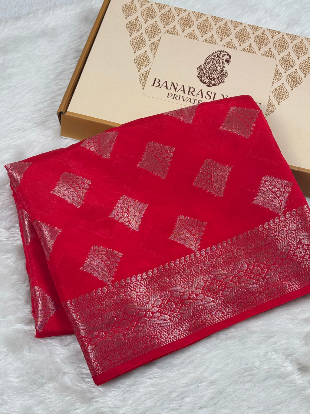 A folded red Banarasi silk saree with gold zari motifs and border, displayed next to its beige packaging box on a white faux fur surface.