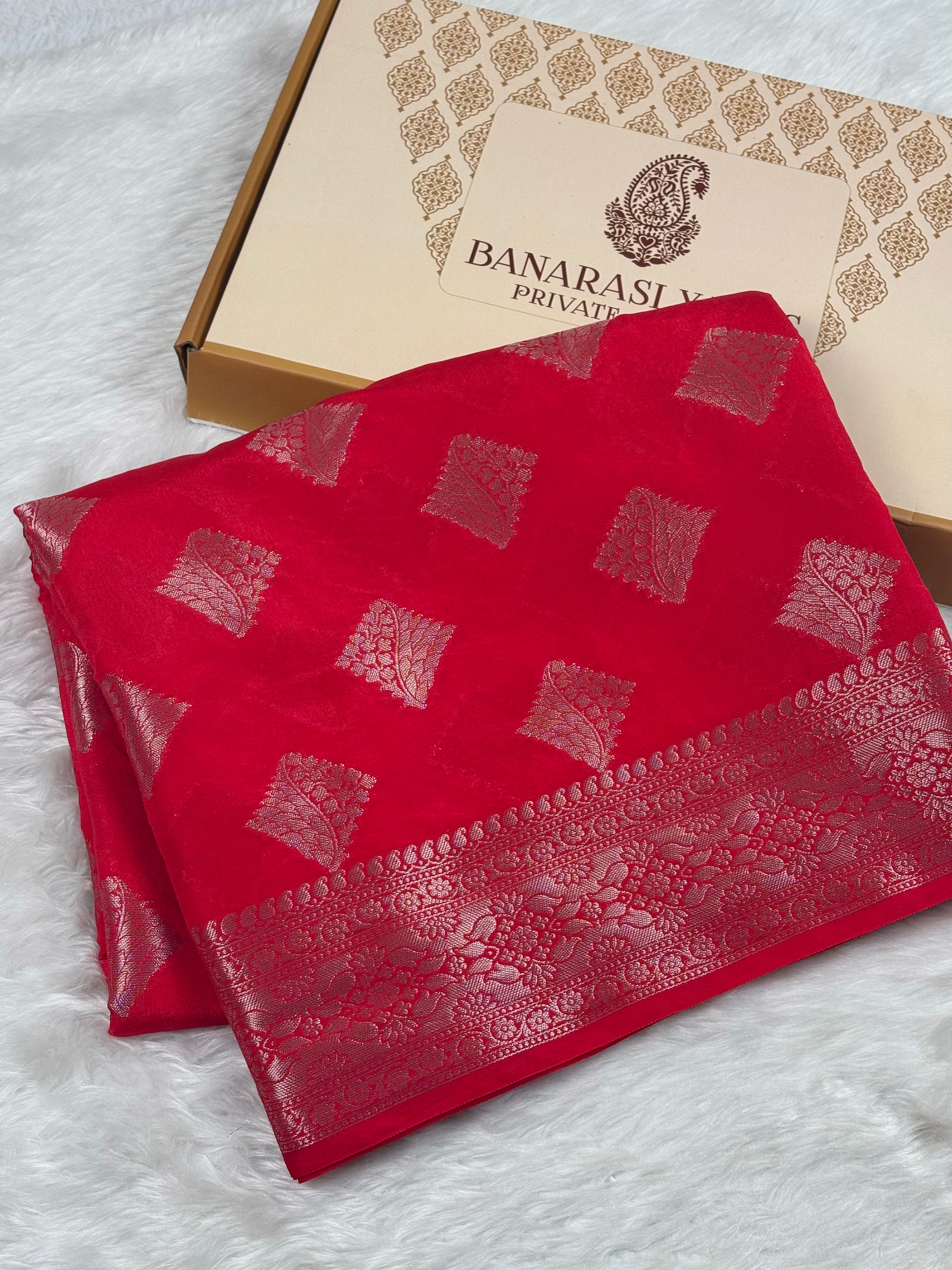A folded red Banarasi silk saree with gold zari motifs and border, displayed next to its beige packaging box on a white faux fur surface.