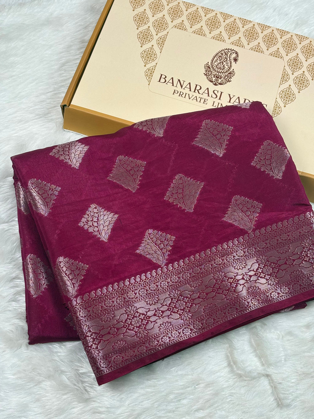 A folded deep magenta Banarasi silk saree with silver zari woven geometric patterns and a wide decorative border, displayed next to its Banarasi Yards Private Limited gift box on a white faux fur surface.