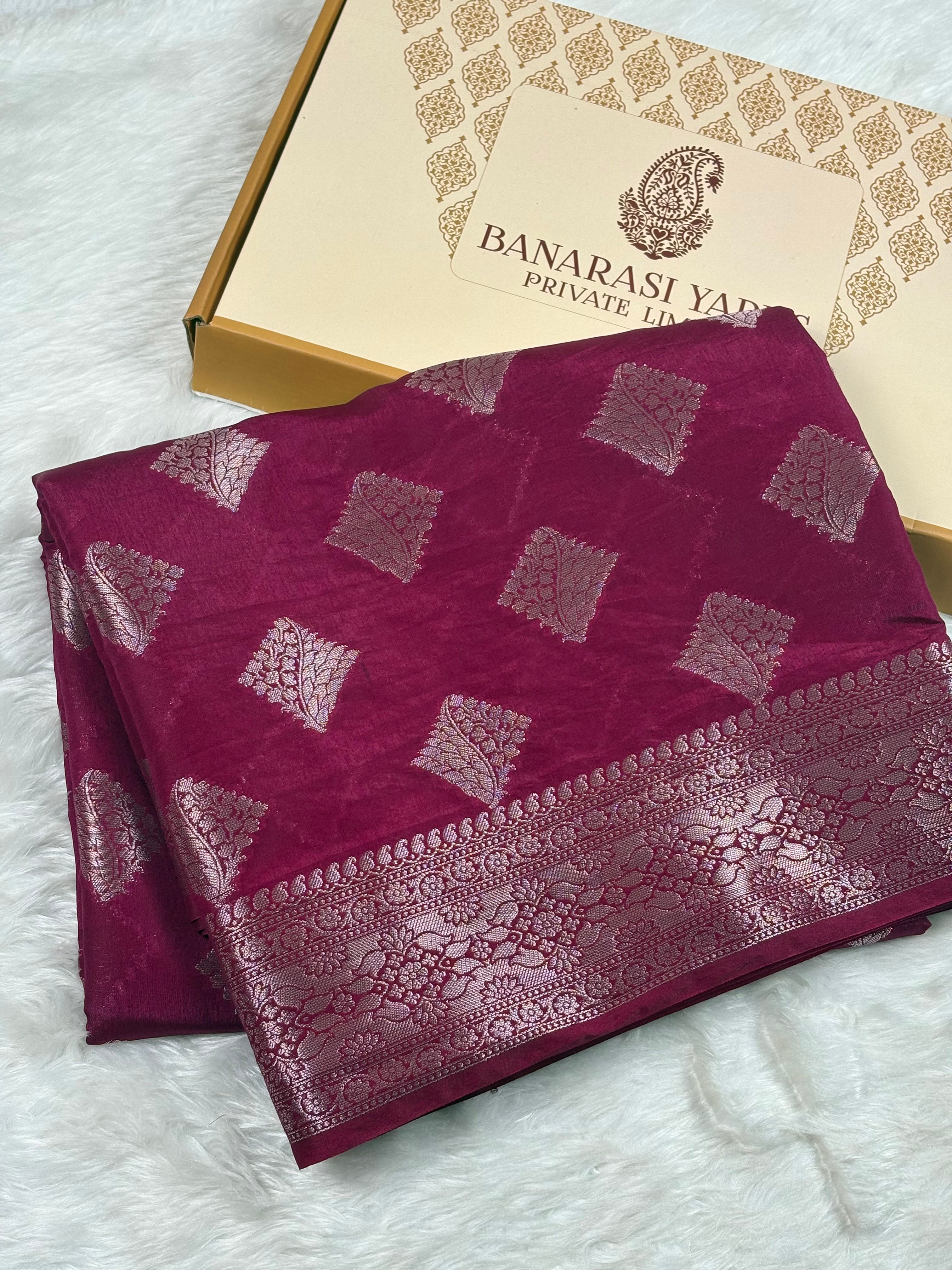 A folded deep magenta Banarasi silk saree with silver zari woven geometric patterns and a wide decorative border, displayed next to its Banarasi Yards Private Limited gift box on a white faux fur surface.