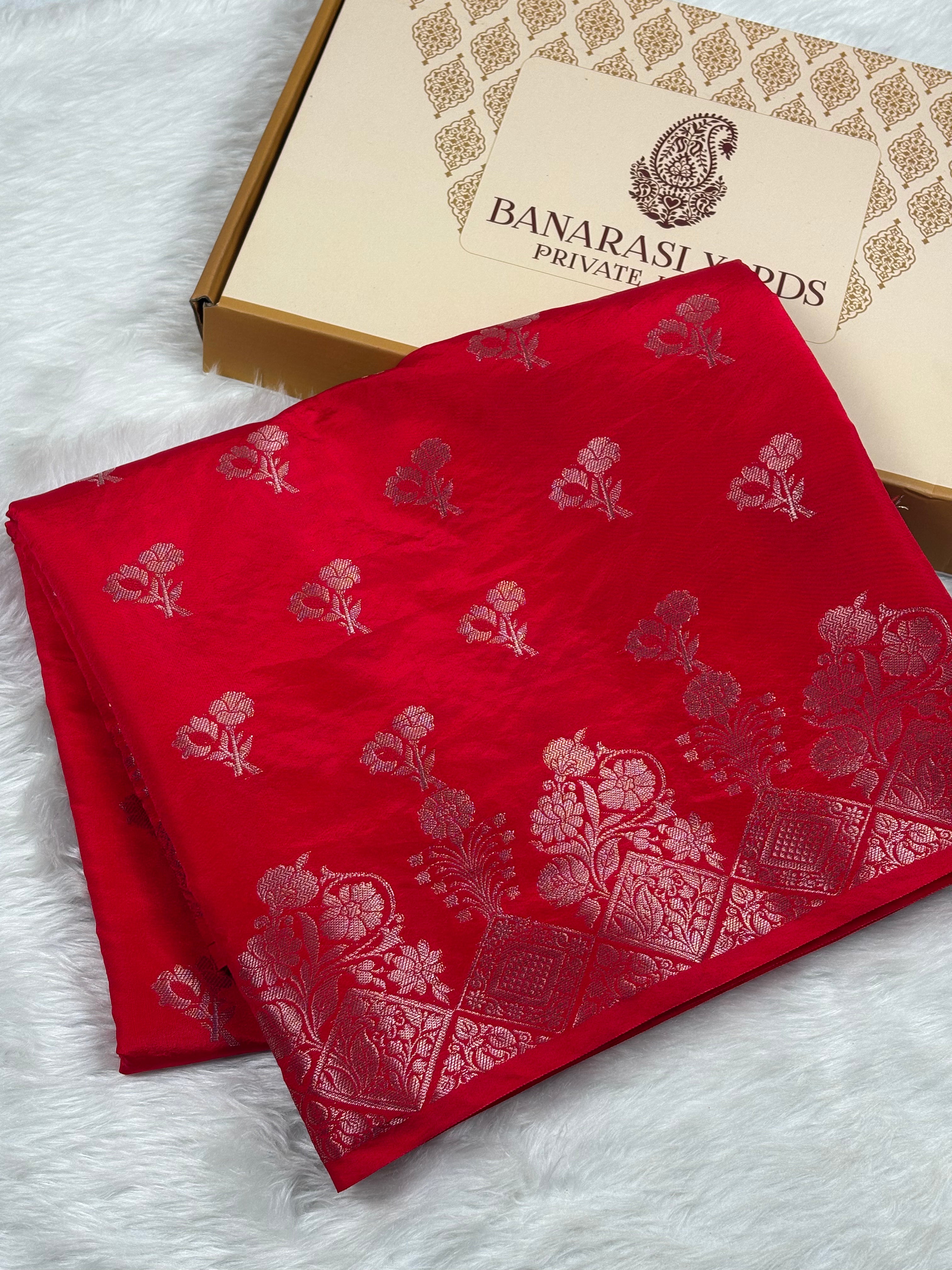 A folded red silk saree with gold floral mina buti and border patterns, displayed next to its Banarasi Yards Private Limited box on a white faux fur surface.