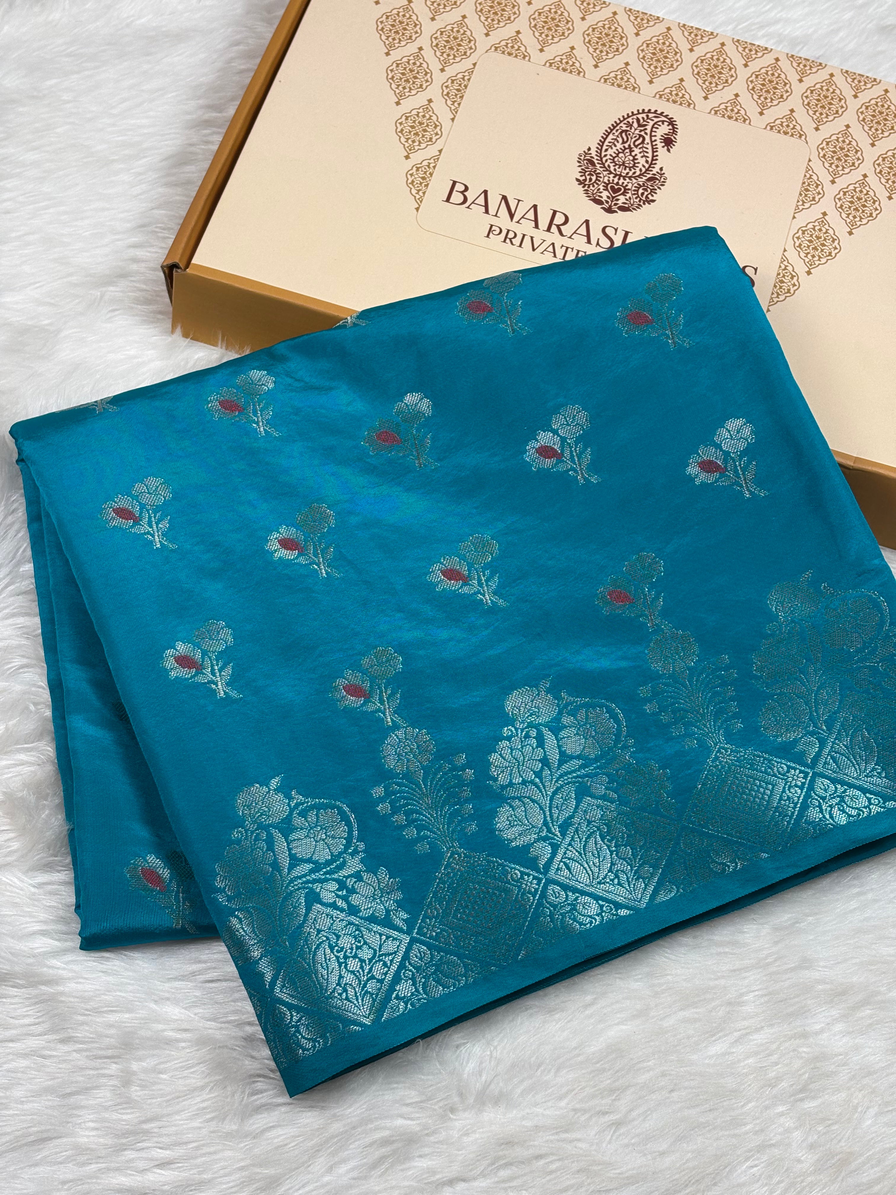 A folded teal blue Banarasi silk saree with intricate silver floral and paisley zari weaving, displayed next to its branded gift box on a white faux fur surface.