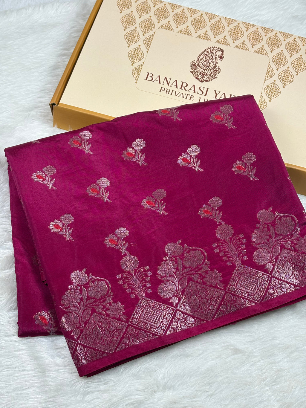 A folded magenta pink Banarasi silk saree with silver floral and paisley zari motifs, displayed next to its branded packaging box on a white faux fur surface.