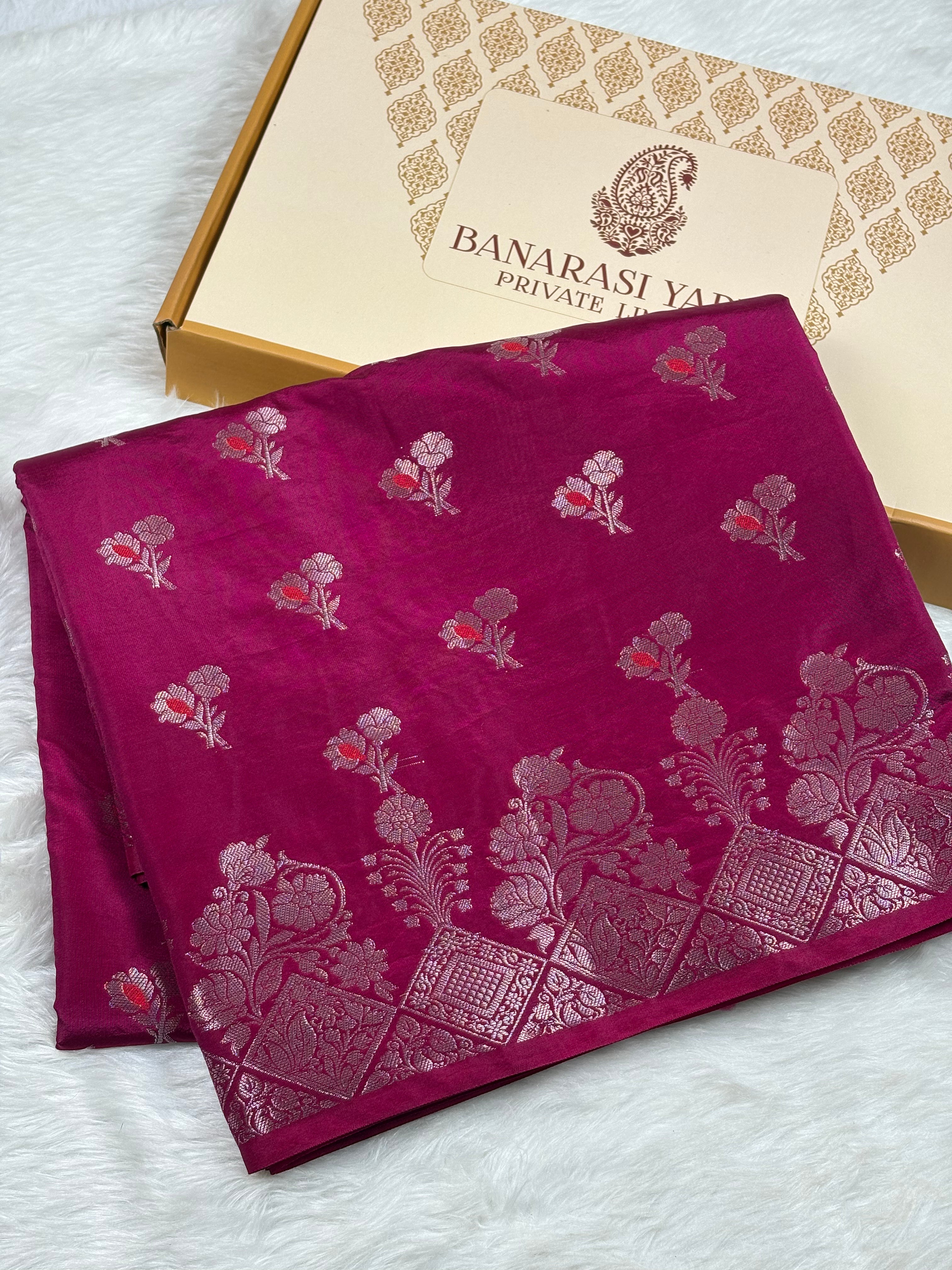 A folded magenta pink Banarasi silk saree with silver floral and paisley zari motifs, displayed next to its branded packaging box on a white faux fur surface.