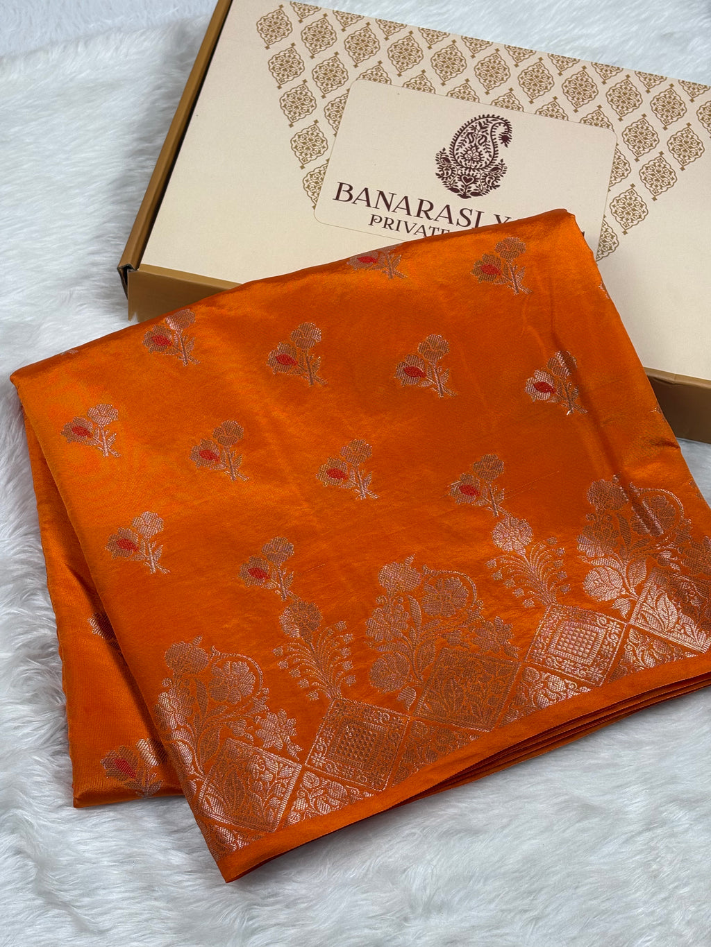An orange silk saree with mina buti floral motifs, displayed next to its branded packaging box on a white faux fur surface.