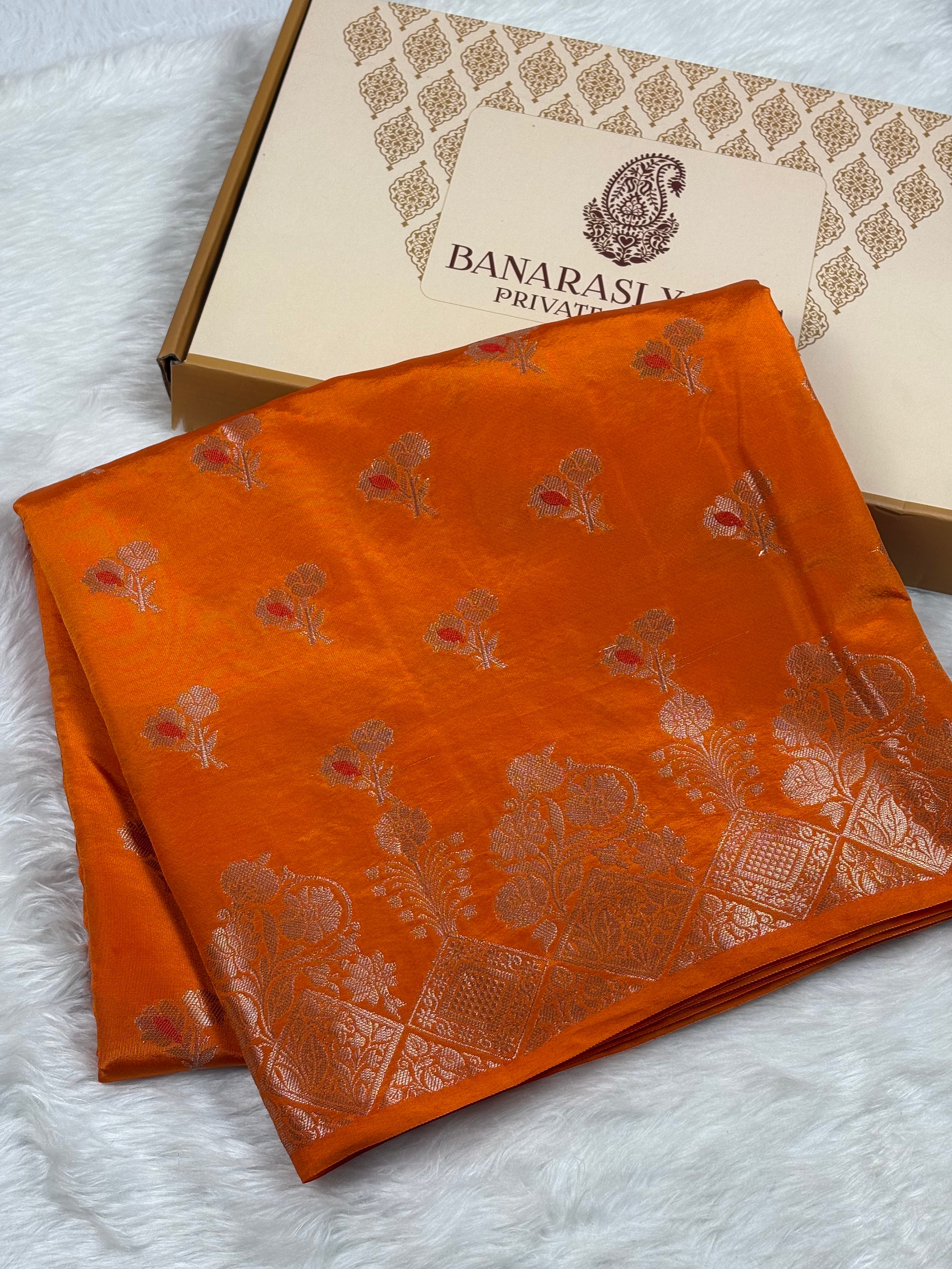 An orange silk saree with mina buti floral motifs, displayed next to its branded packaging box on a white faux fur surface.