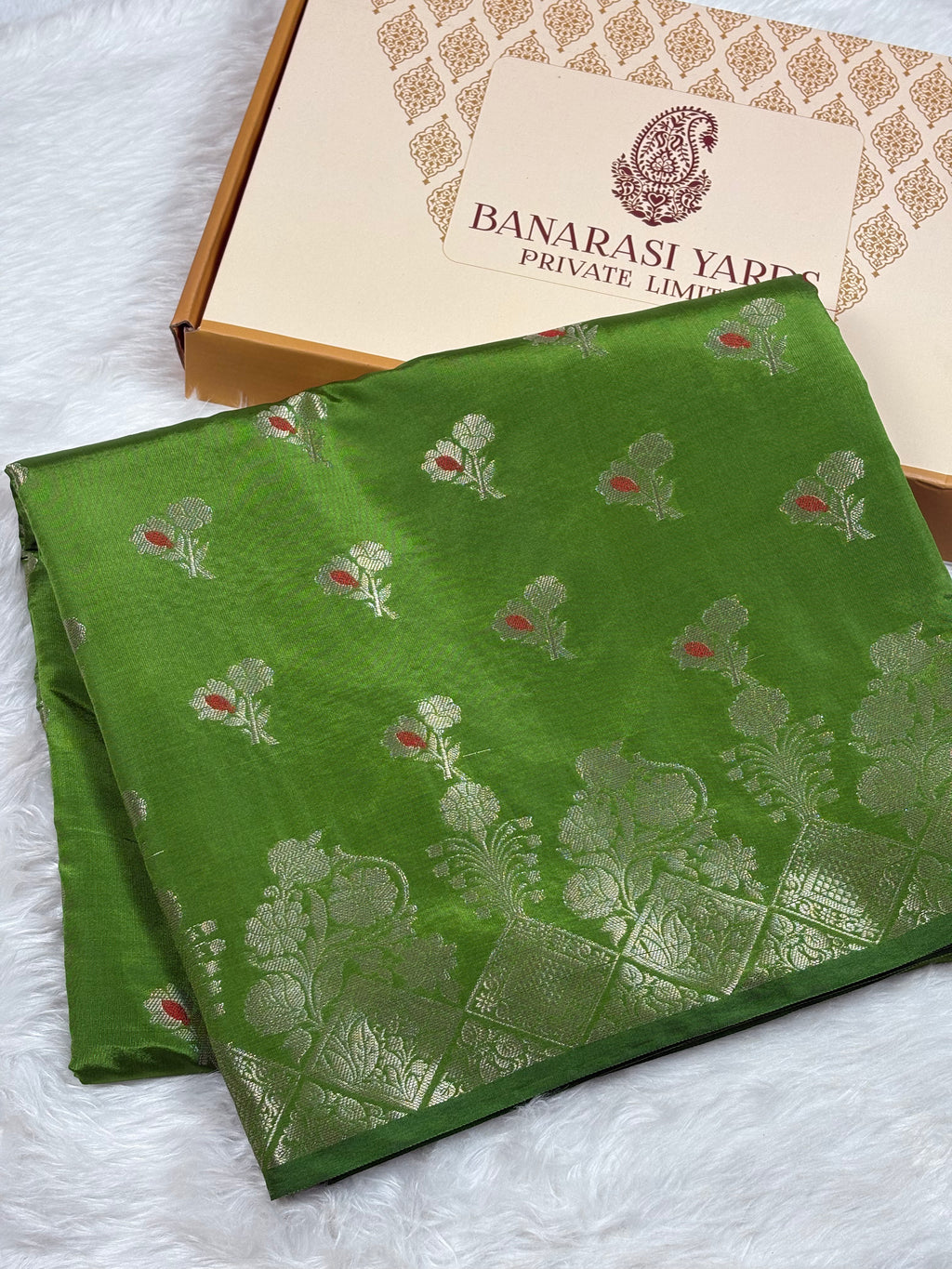 A folded green silk saree with silver floral motifs and small mina accents, displayed next to its Banarasi Yards box on a white faux fur surface.